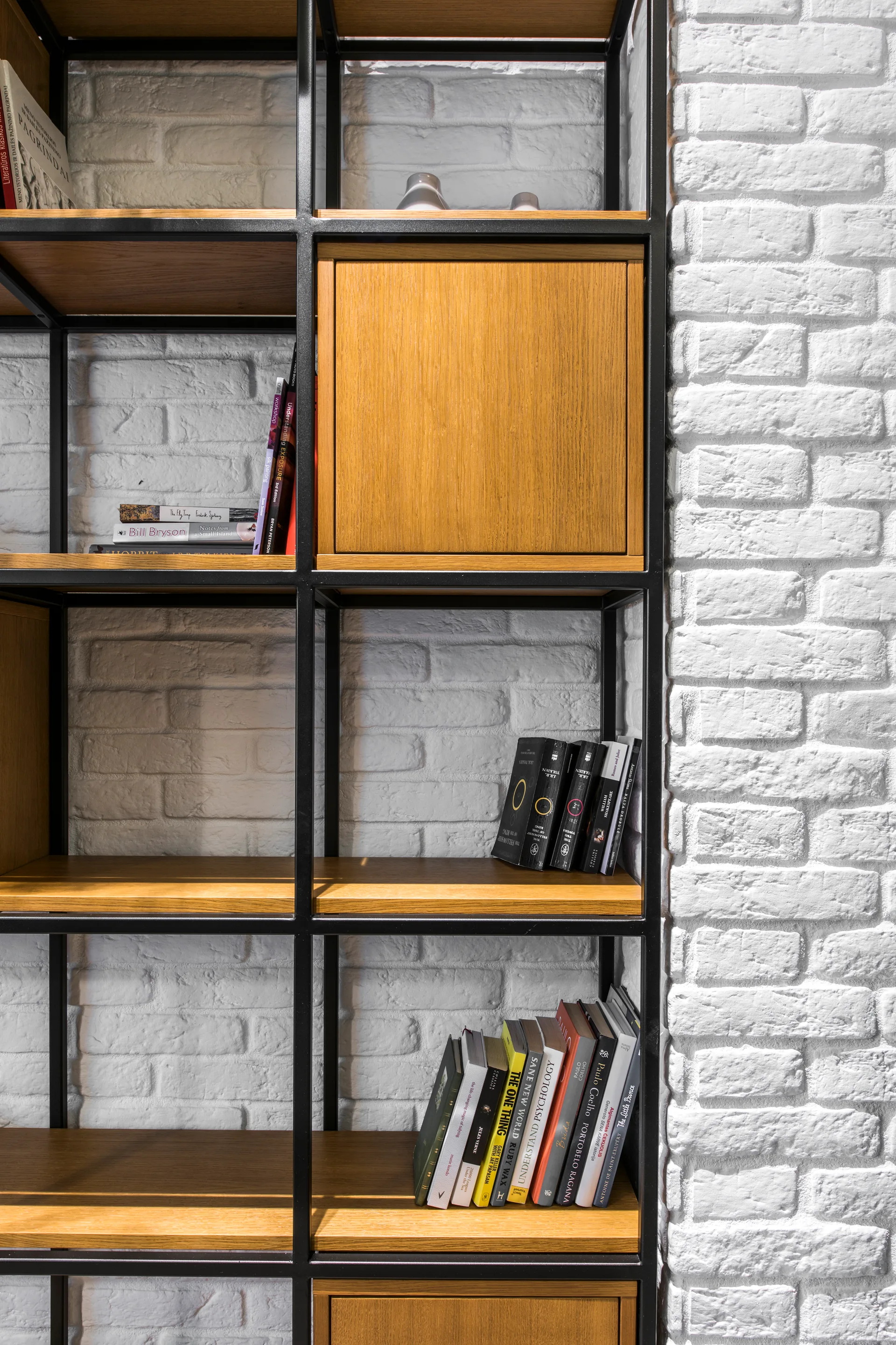 A black metal grid bookshelf with warm wood shelves and integrated wood cabinets stands in front of a white painted brick wall.