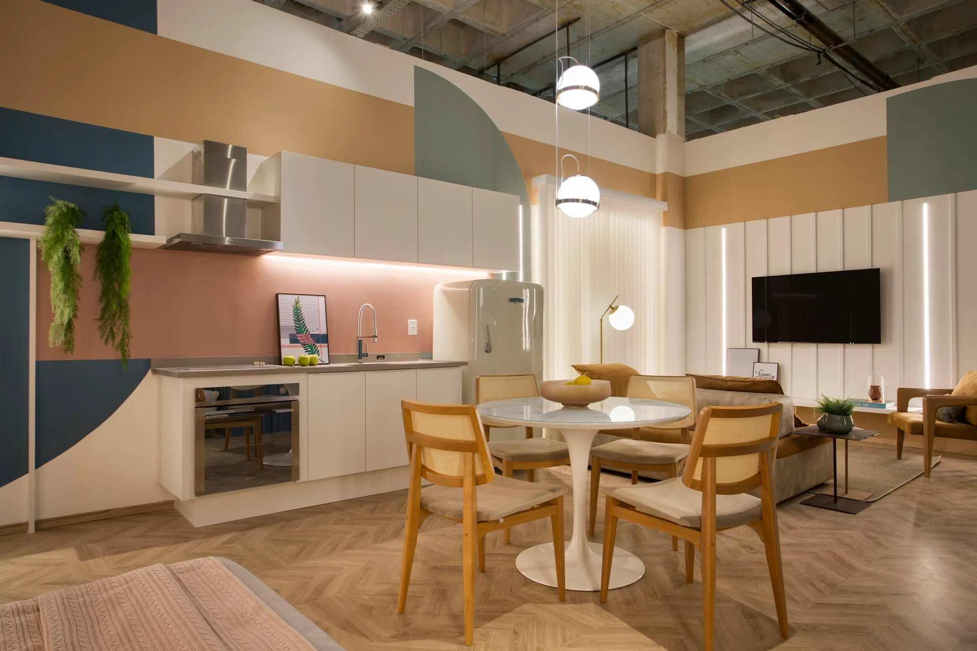 A contemporary studio interior with a white kitchen, a round dining set, chevron wood floors, and a colorful painted geometric accent wall.
