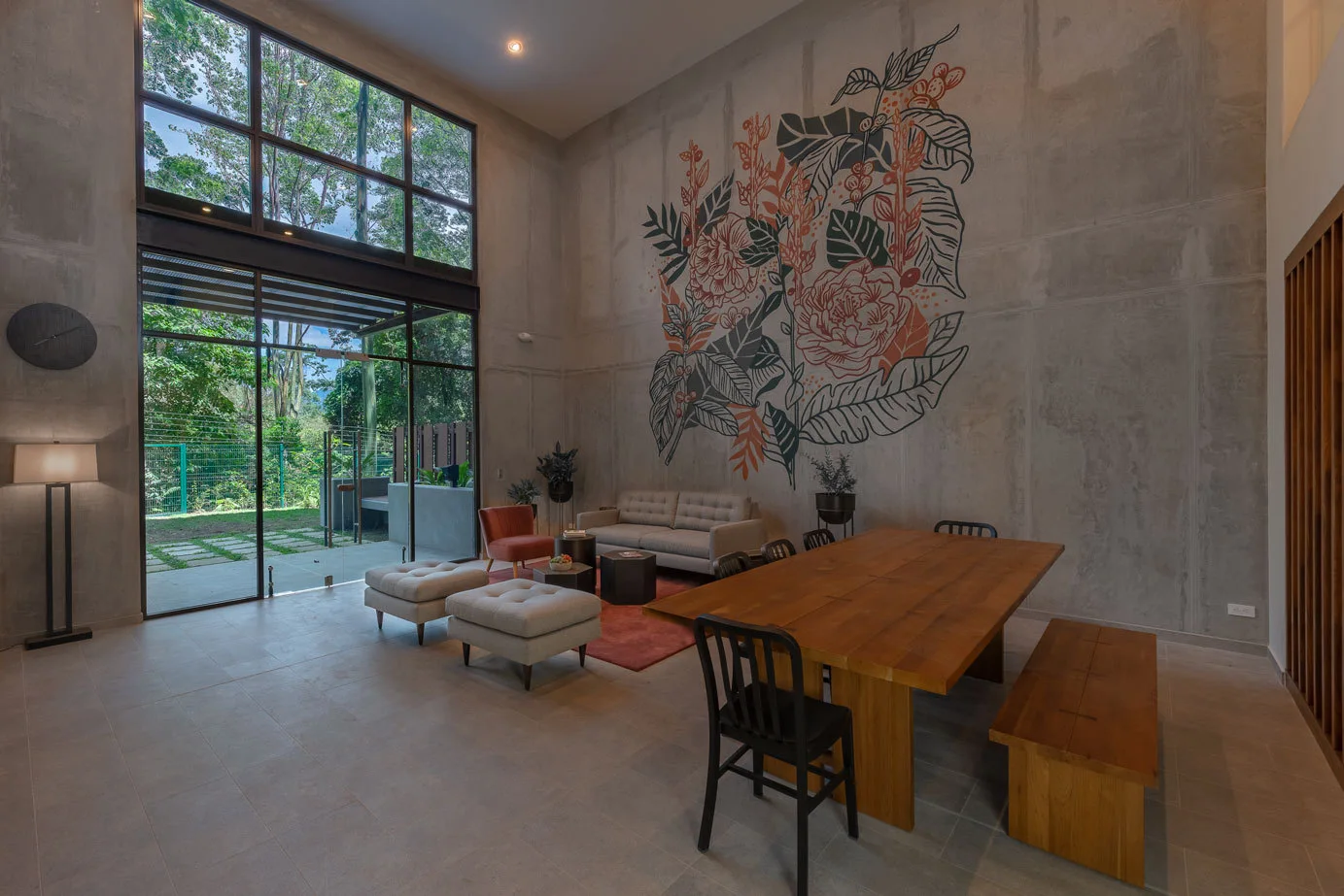 A double-height living and dining space features a large botanical mural on an industrial concrete wall alongside a wooden dining table and a neutral sofa.