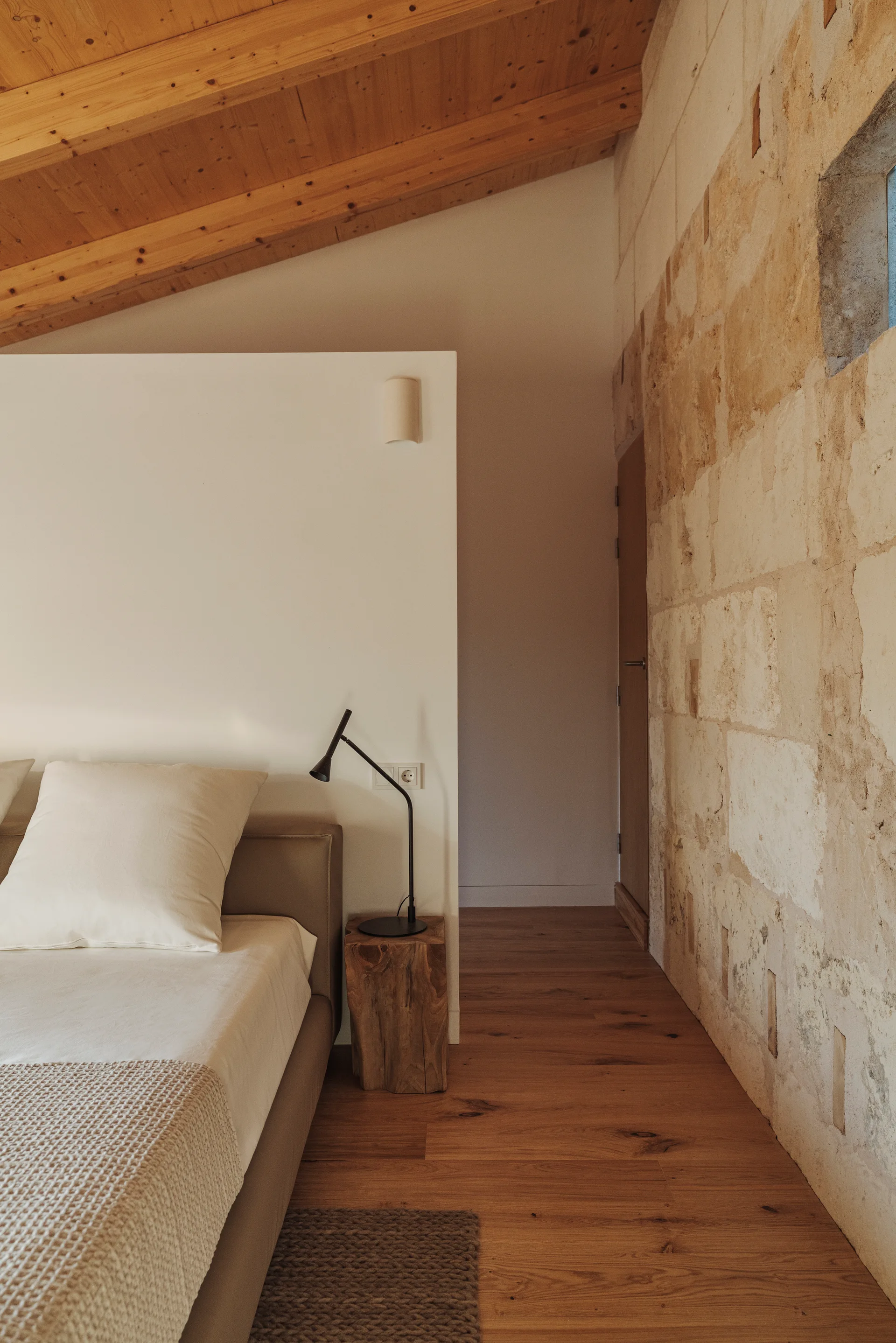 A minimalist bedroom featuring an upholstered bed, a rustic wood block nightstand, an exposed stone wall, and a sloped wood ceiling.