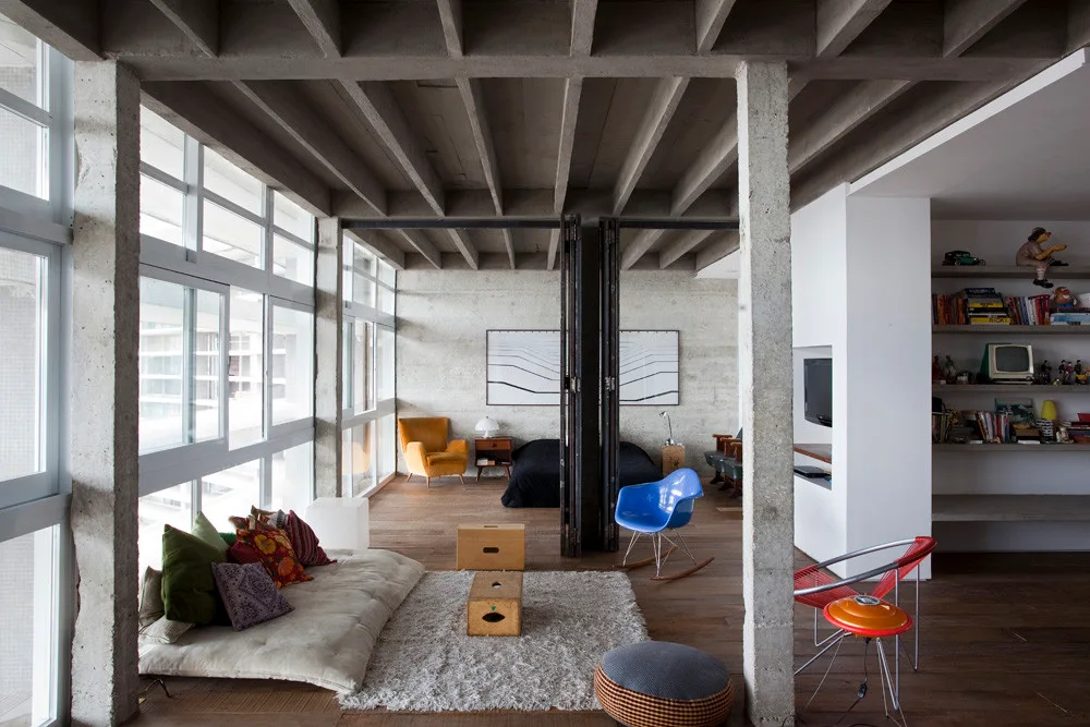 An open-plan loft featuring an exposed concrete waffle ceiling, concrete columns, wood flooring, a low-profile floor sofa, and a blue shell rocking chair.