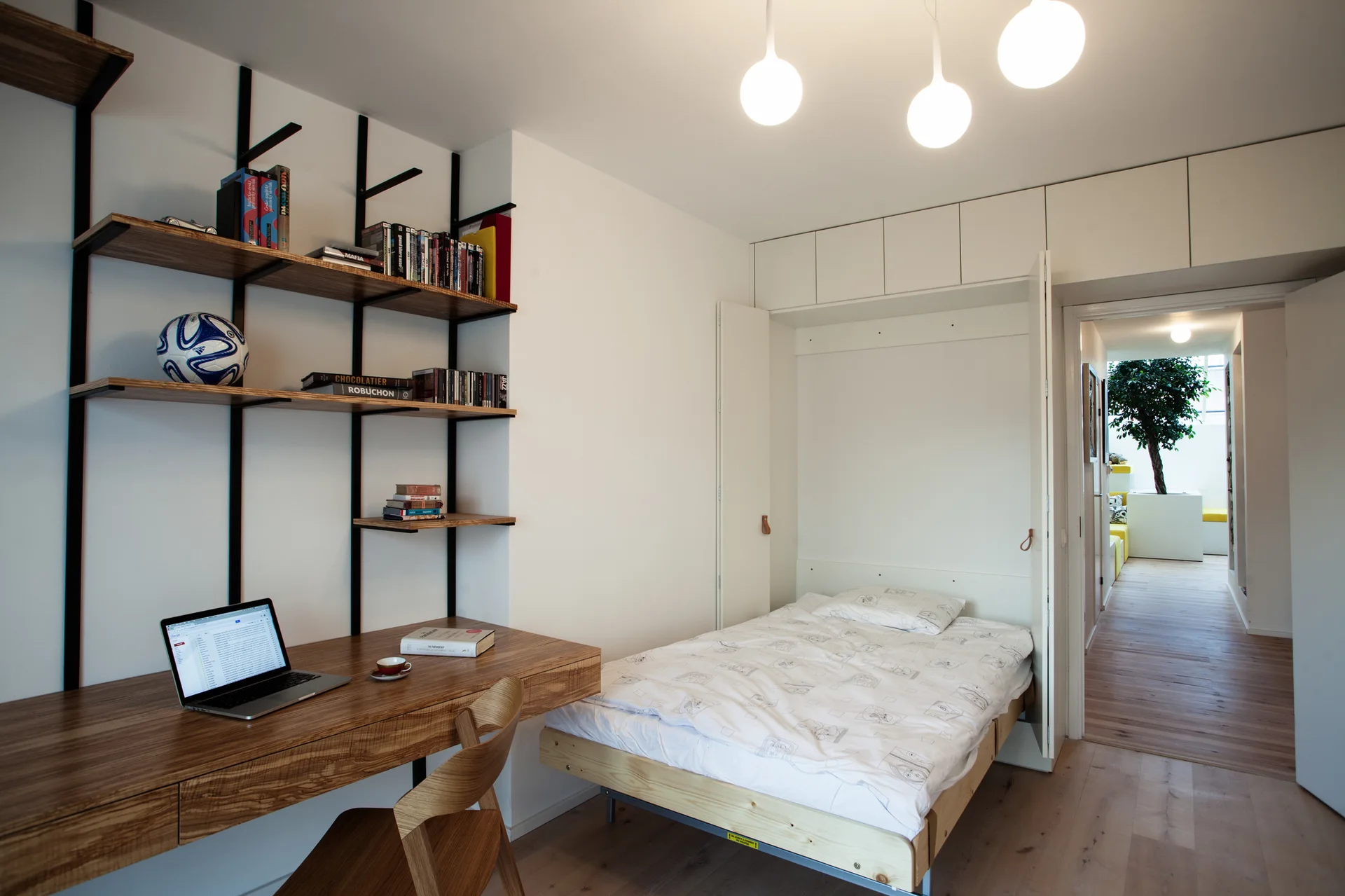 A compact modern bedroom featuring a wood desk with shelving, a white wall-integrated Murphy bed, and light wood floors.