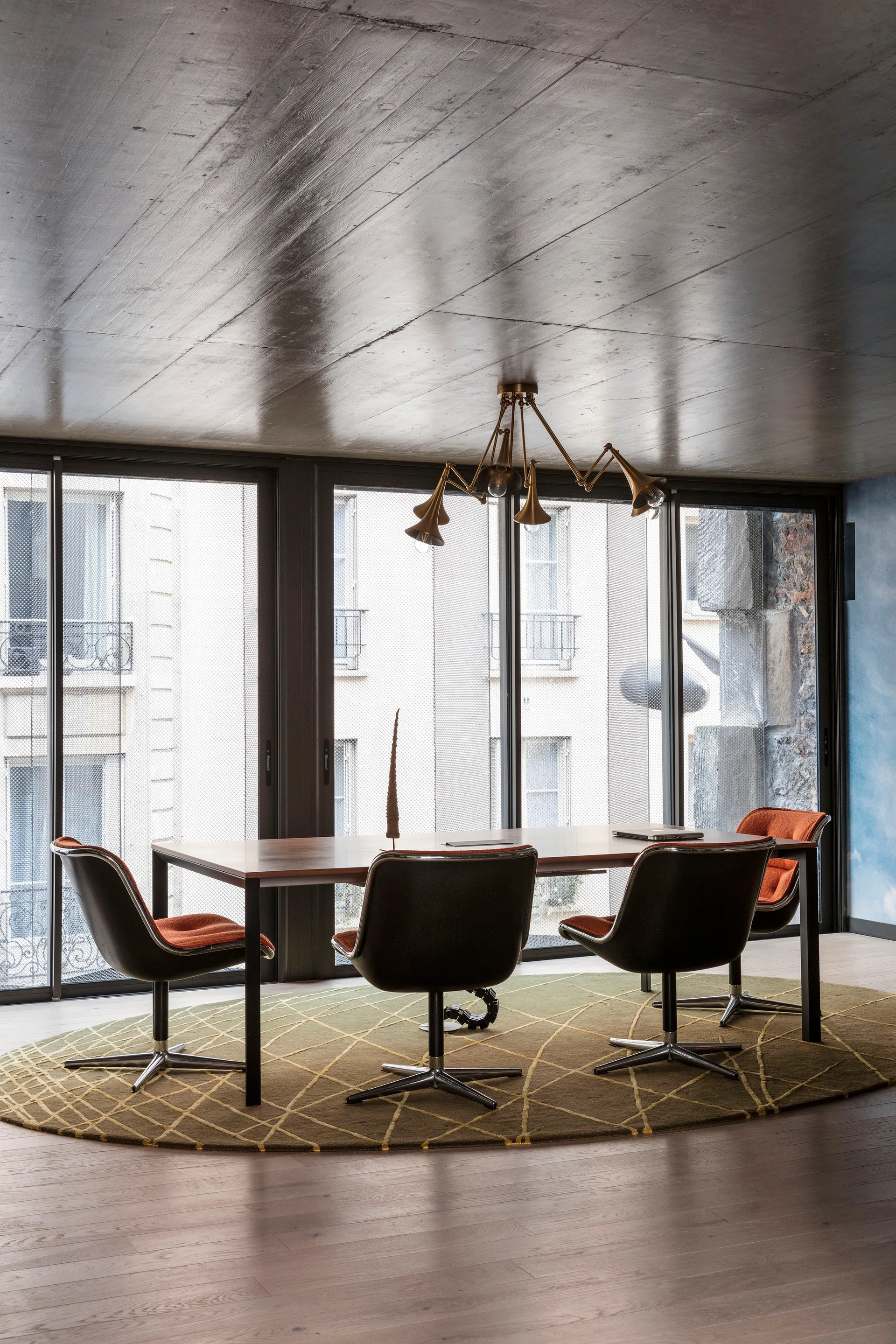 A contemporary dining room featuring a wooden table, swivel chairs with orange cushions, a brass chandelier, and an oval rug in front of large windows.