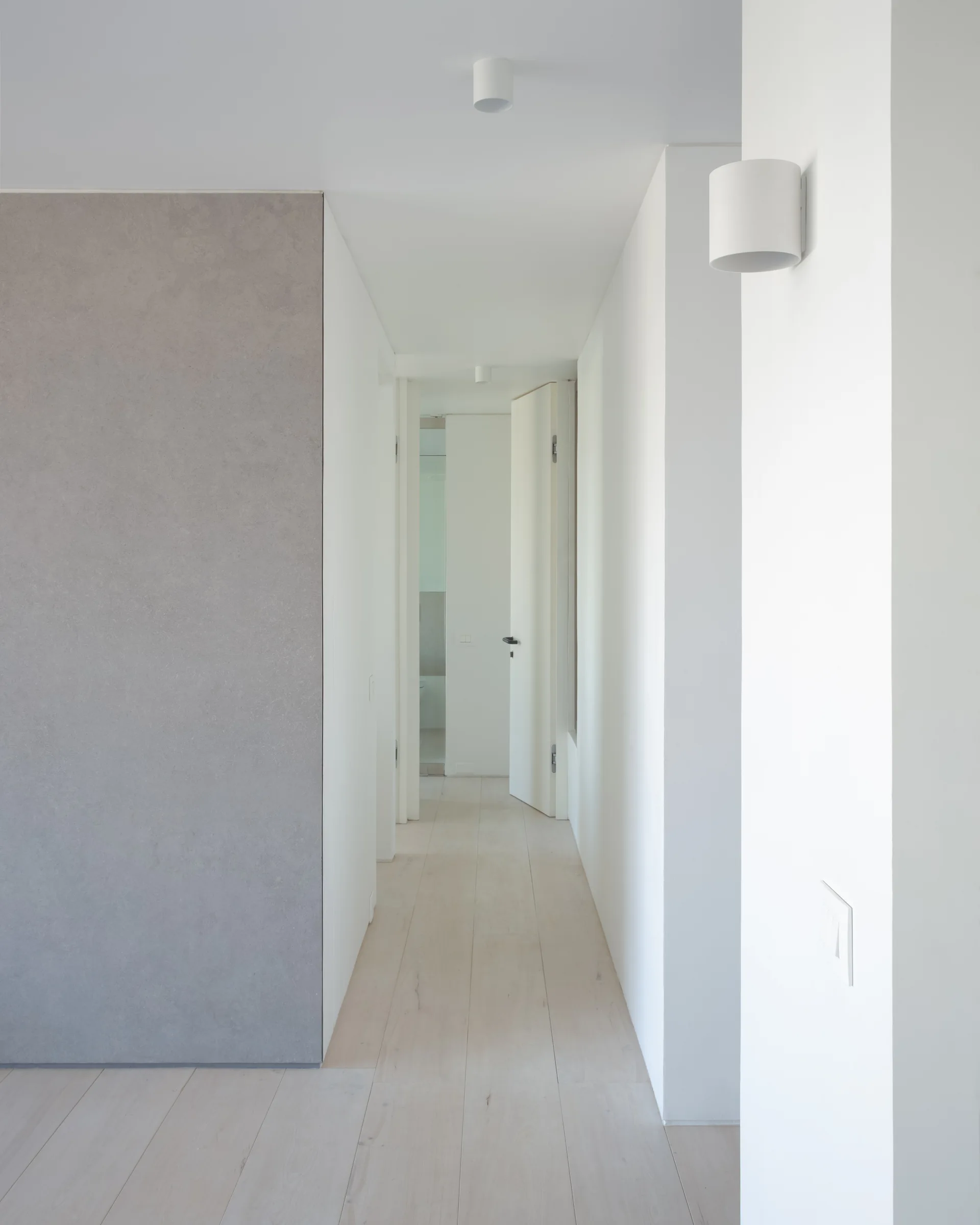 A minimalist hallway features a matte grey accent wall, white walls, light wood plank floors, and matching white cylindrical sconces.