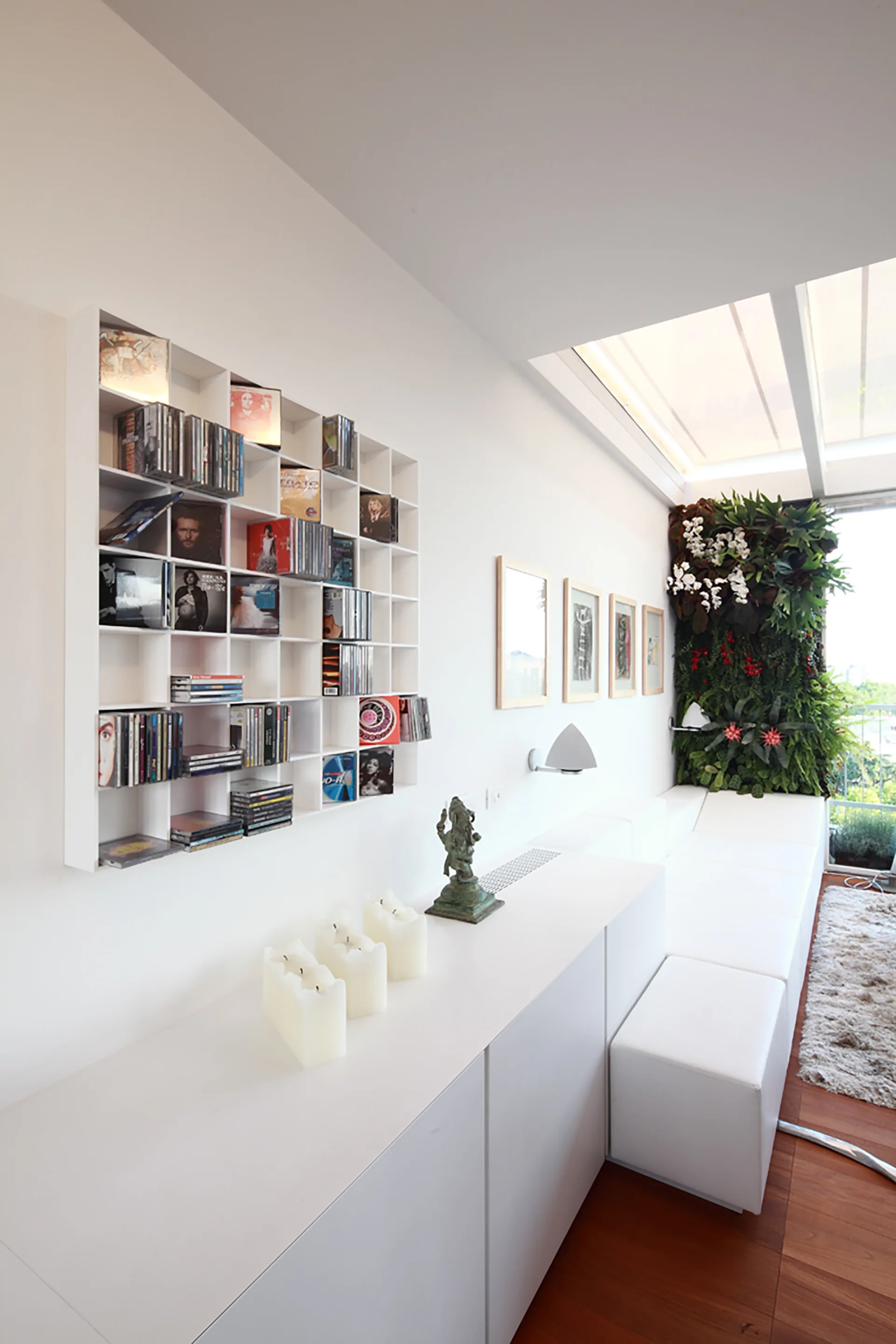 A minimalist white living room features a grid wall shelf, low-profile white cabinets, modular seating, and a lush vertical green garden under a skylight.