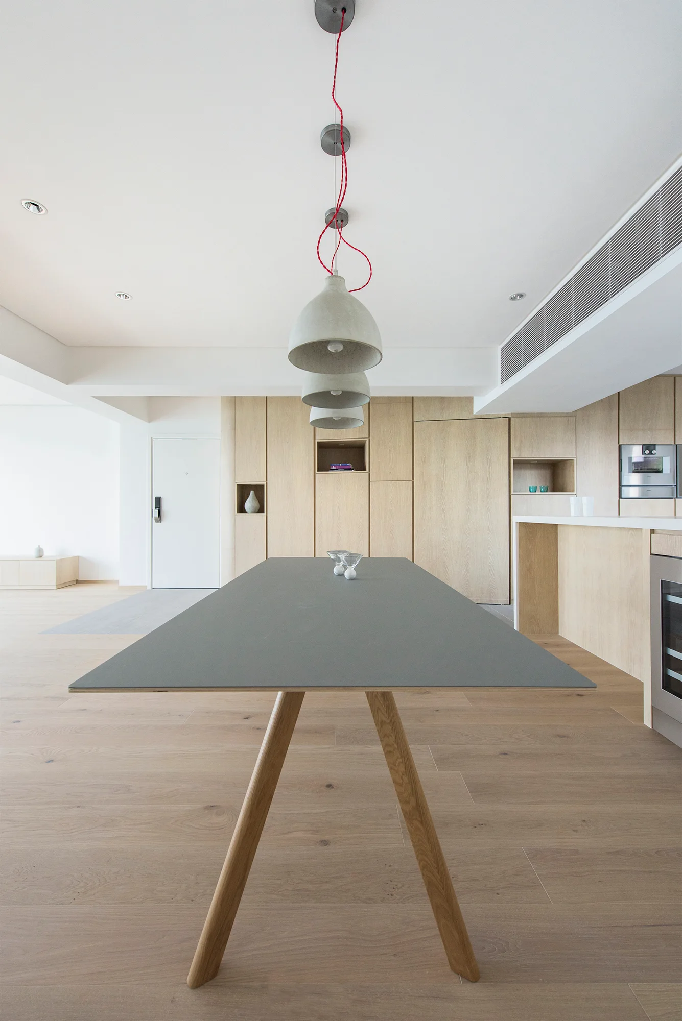 A minimalist grey dining table with wood legs sits on light oak floors beneath three concrete pendant lights with red cords.