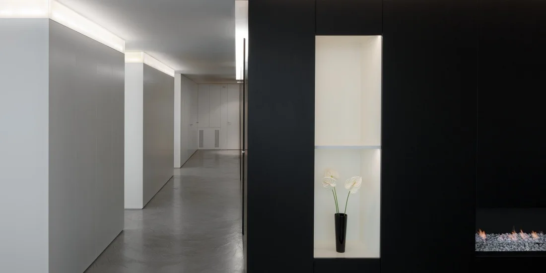 A minimalist hallway features matte black built-in cabinets with a white recessed niche containing a vase, set against white walls with cove lighting.