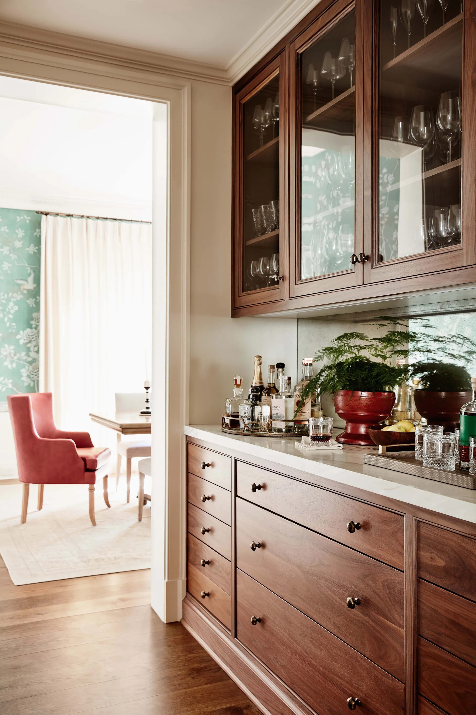 A custom walnut home bar features glass-front upper cabinets, a mirrored backsplash, and marble countertops, looking into a dining room with a pink armchair.