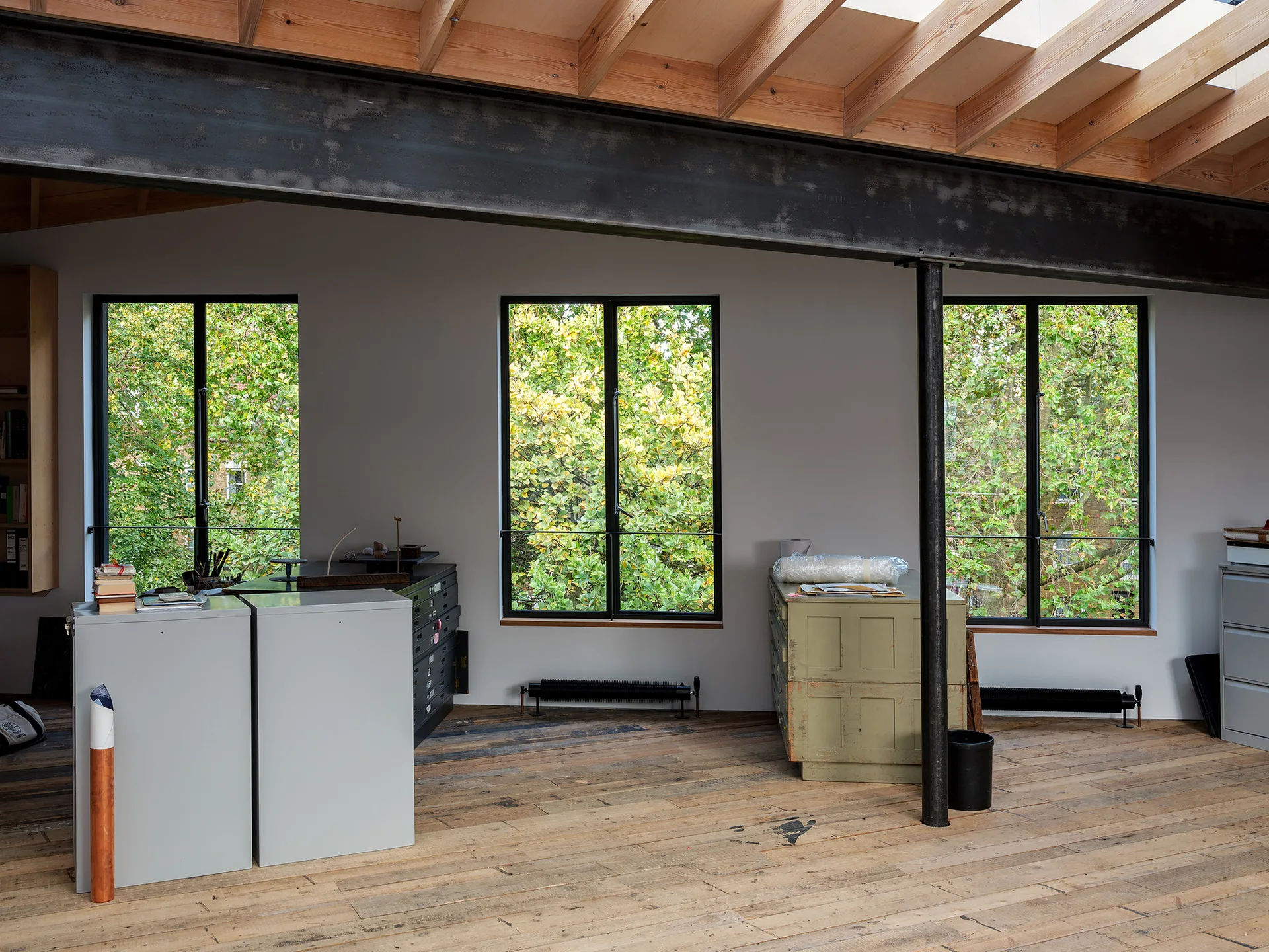 An industrial office features three black-framed windows overlooking green trees, exposed wood rafters, a massive steel beam, and rustic wood flooring.