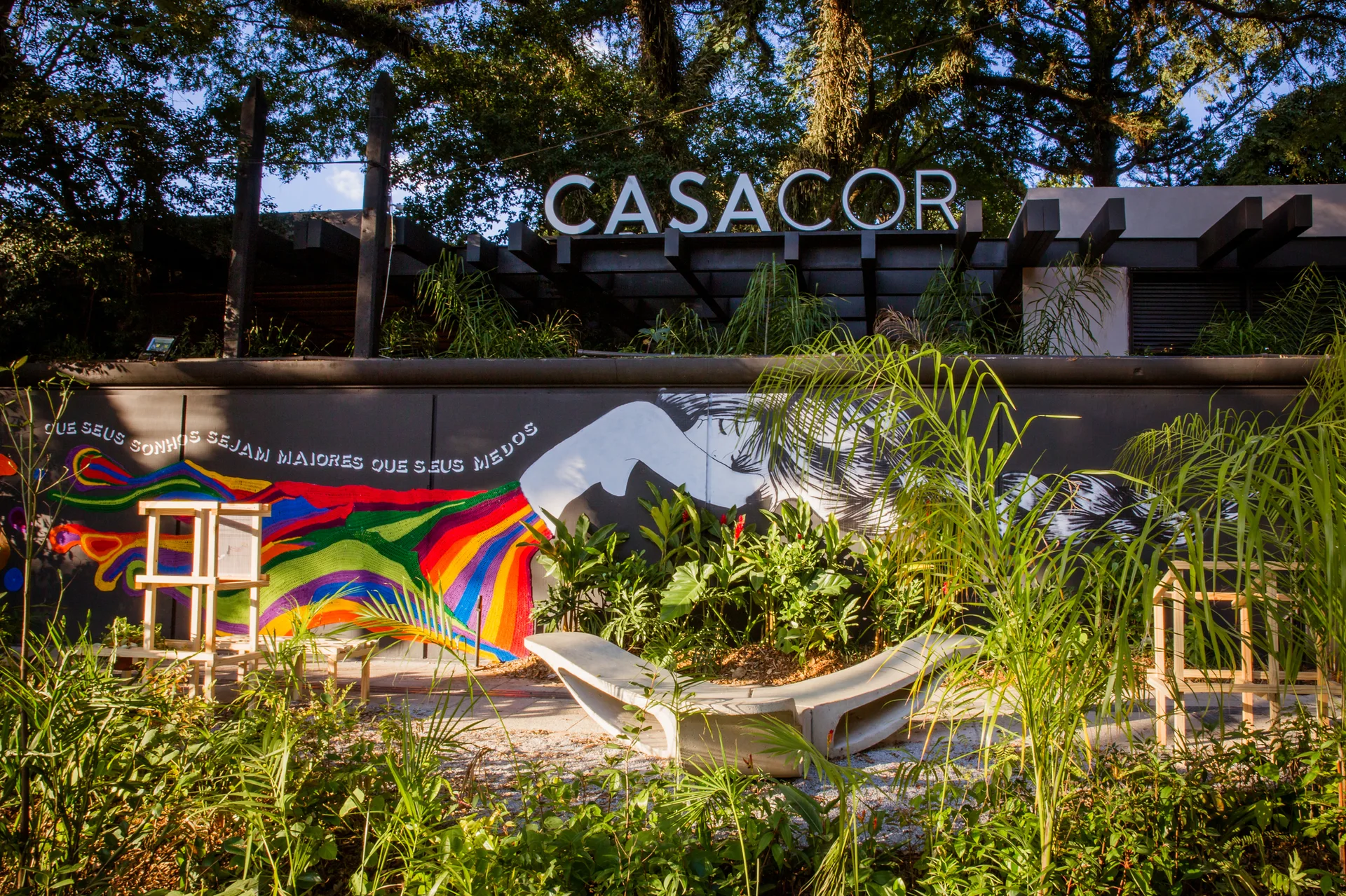 A curved concrete bench sits in an outdoor garden courtyard in front of a dark wall featuring a multicolored graphic mural.
