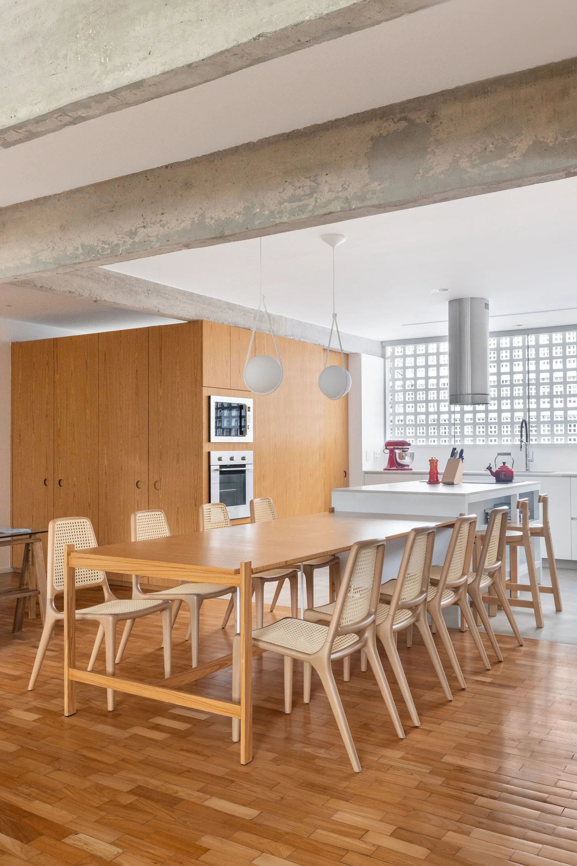 A wooden dining table surrounded by cane-back chairs sits next to a white kitchen island under an exposed concrete beam.
