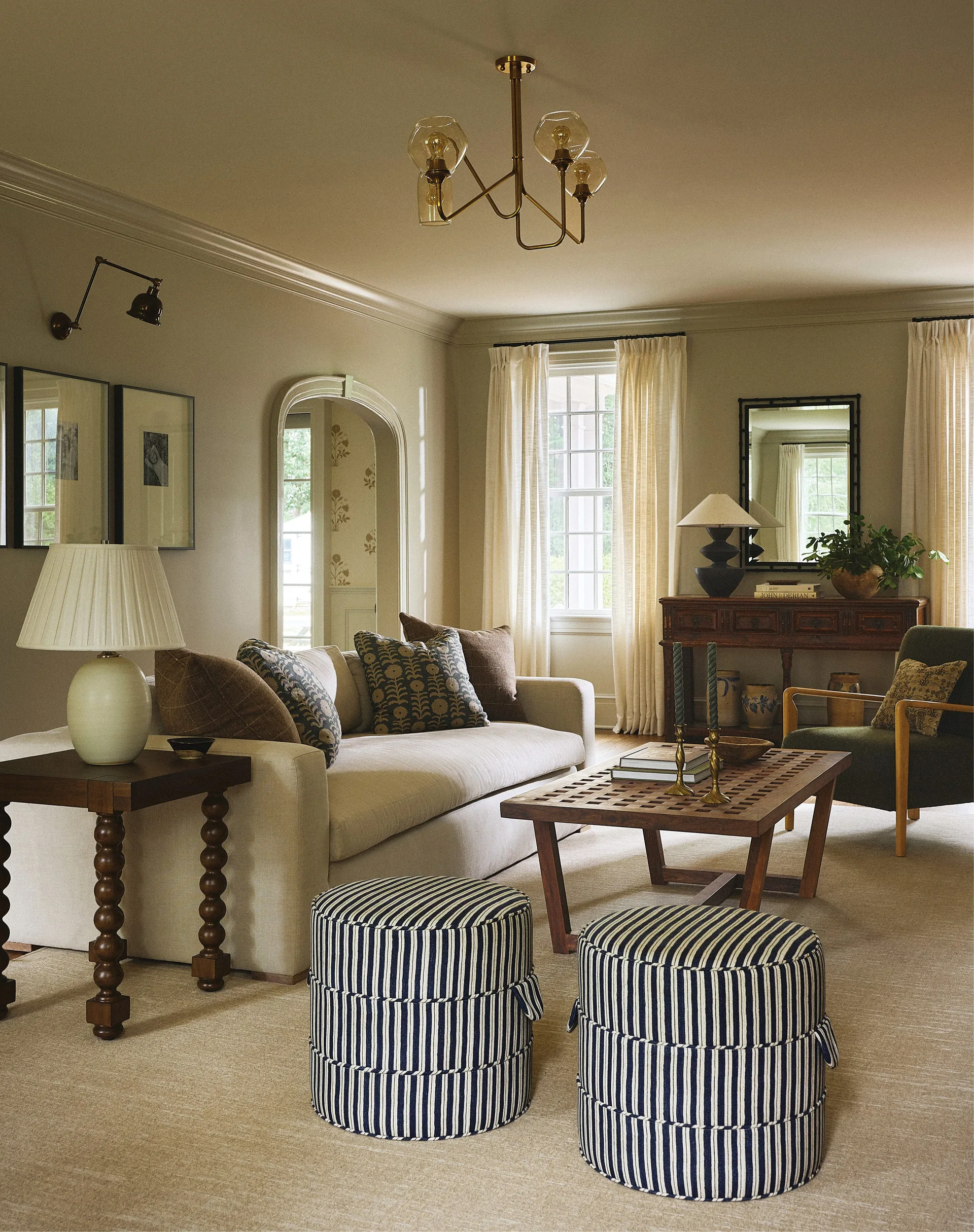 A traditional living room features a beige sofa, two blue and white striped ottomans, a wooden coffee table, and an arched doorway.