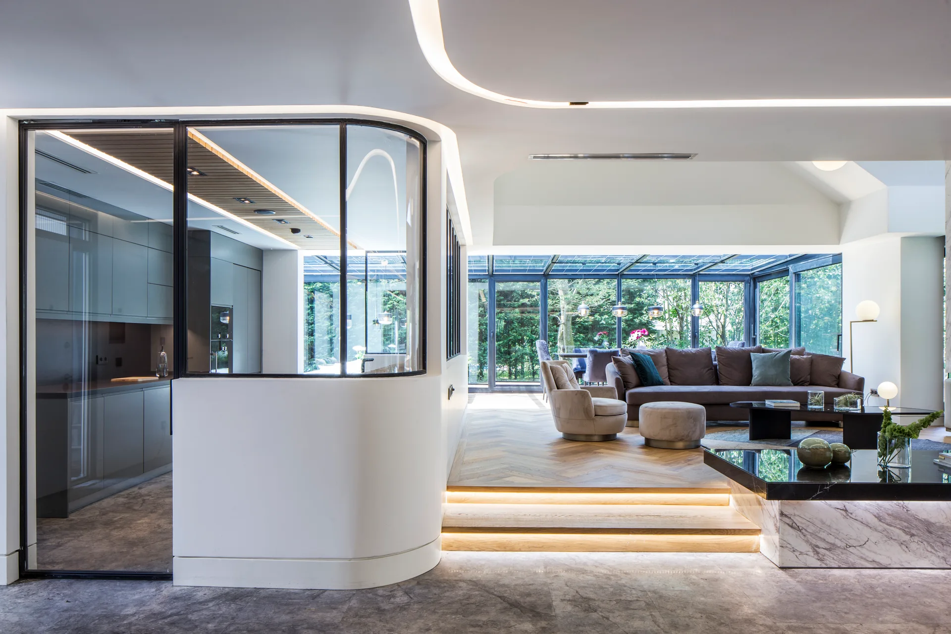 A modern living room features a curved glass partition, wood herringbone flooring, integrated step lighting, a grey sectional sofa, and garden views.
