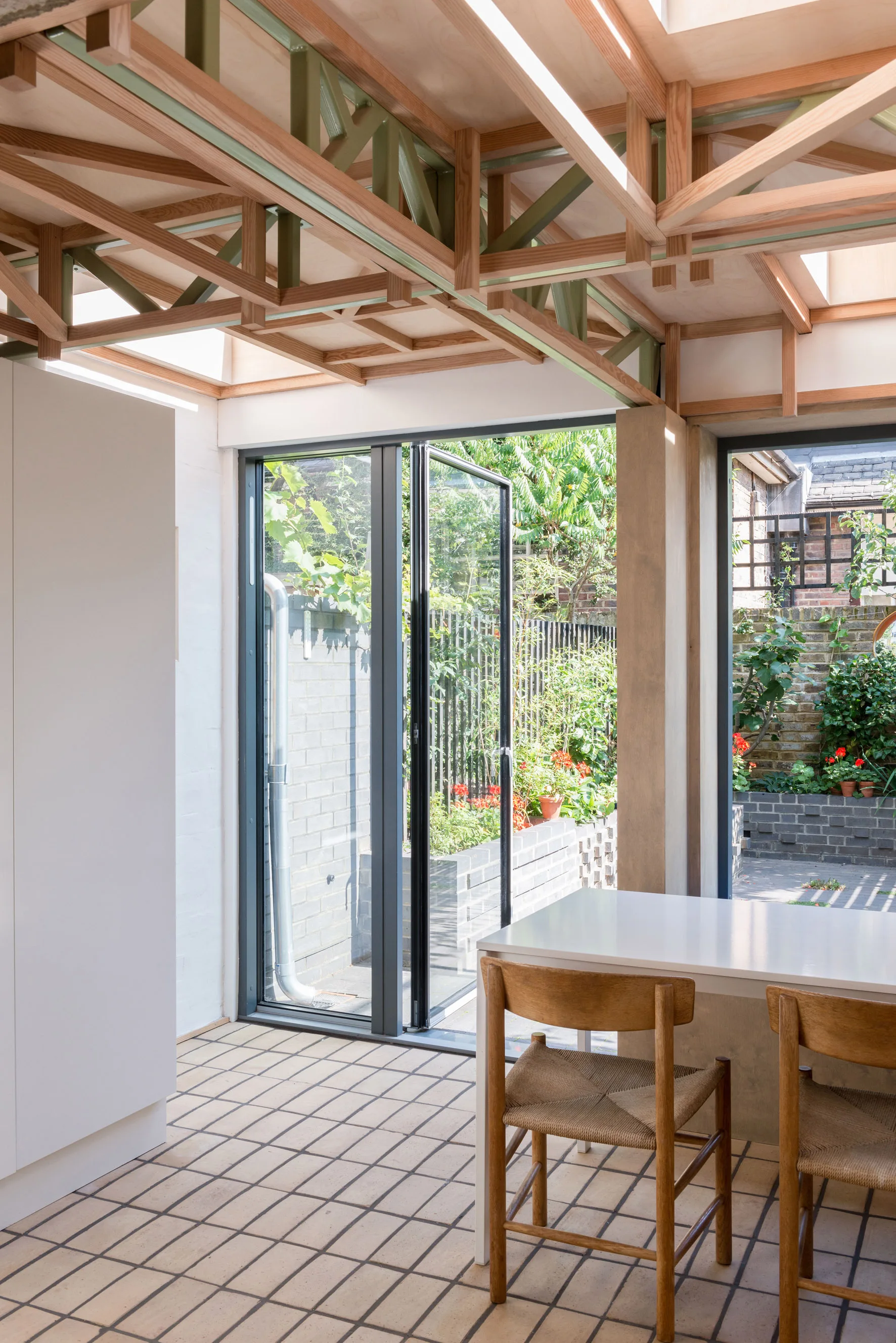 A bright dining room with an exposed wooden grid ceiling, glass bifold doors opening to a garden, and a white table with woven wooden chairs.