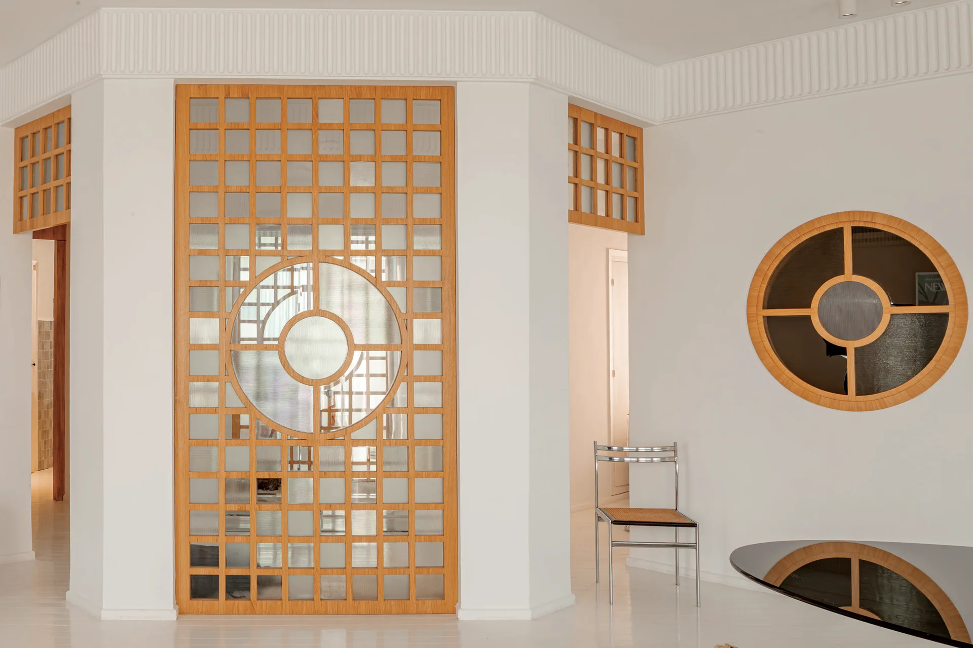 A minimalist white room features a large geometric wood and glass screen divider, a circular window in the wall, and a chrome chair.