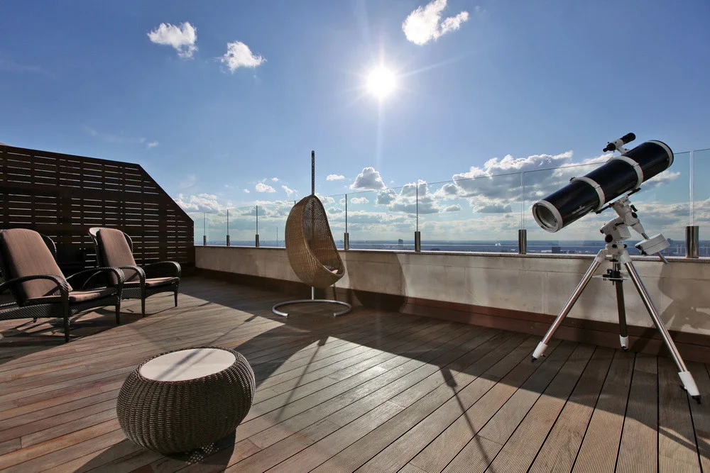 A sunny rooftop terrace featuring wood plank decking, wicker lounge furniture, a hanging egg chair, and a tripod-mounted telescope by a glass railing.