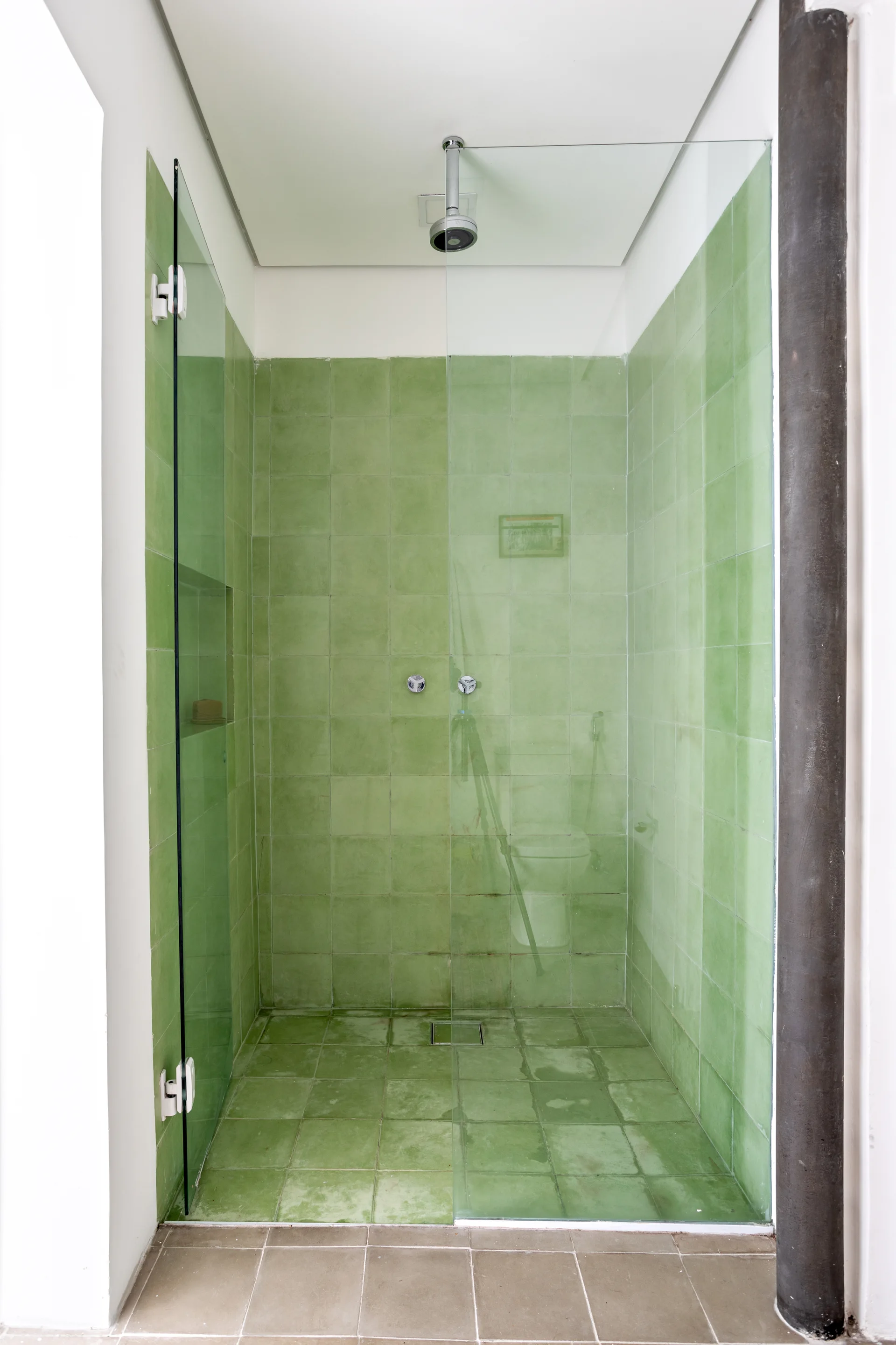 A walk-in shower stall lined with square sage green tiles on the walls and floor, enclosed by a frameless glass partition.