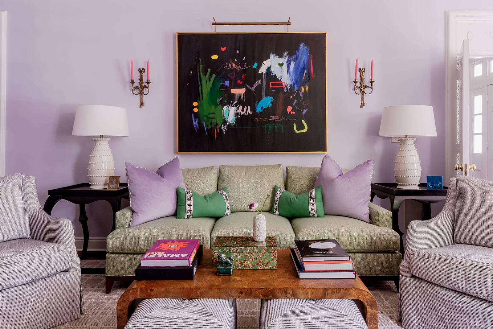 A green sofa with purple pillows sits between white ceramic lamps against a lavender wall with a large abstract painting.