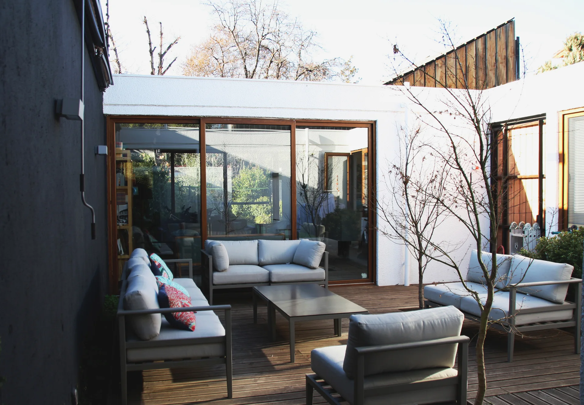 Gray outdoor sofas and armchairs are arranged on a wooden deck next to large sliding glass doors and white stucco walls.