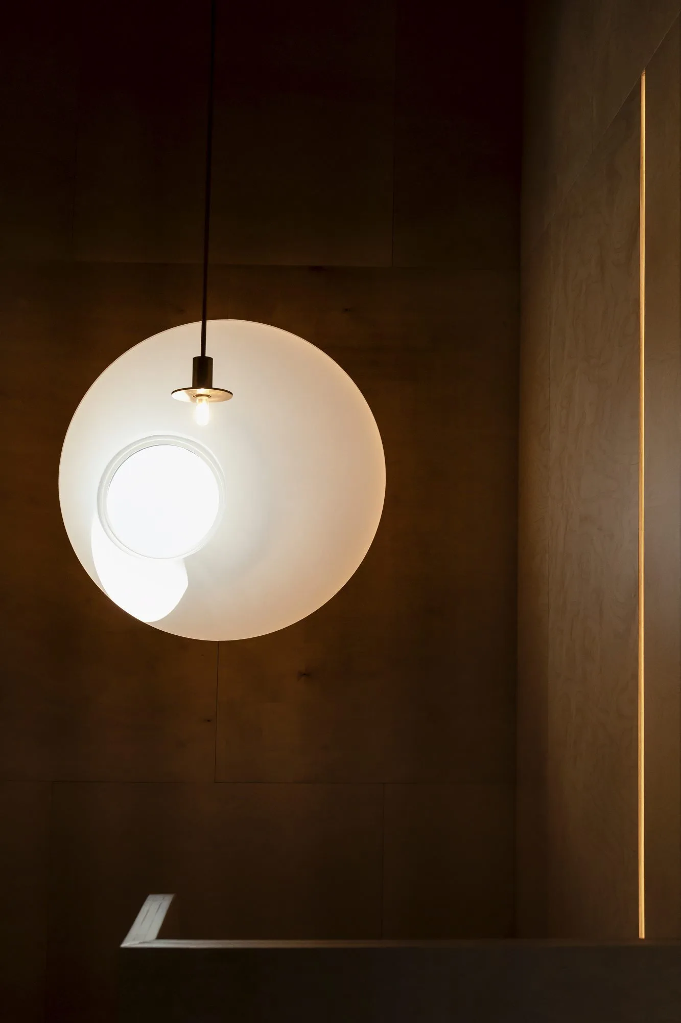 A glowing white circular pendant light hangs in front of dark wood wall paneling, beside a vertical glowing light strip recessed in a seam.