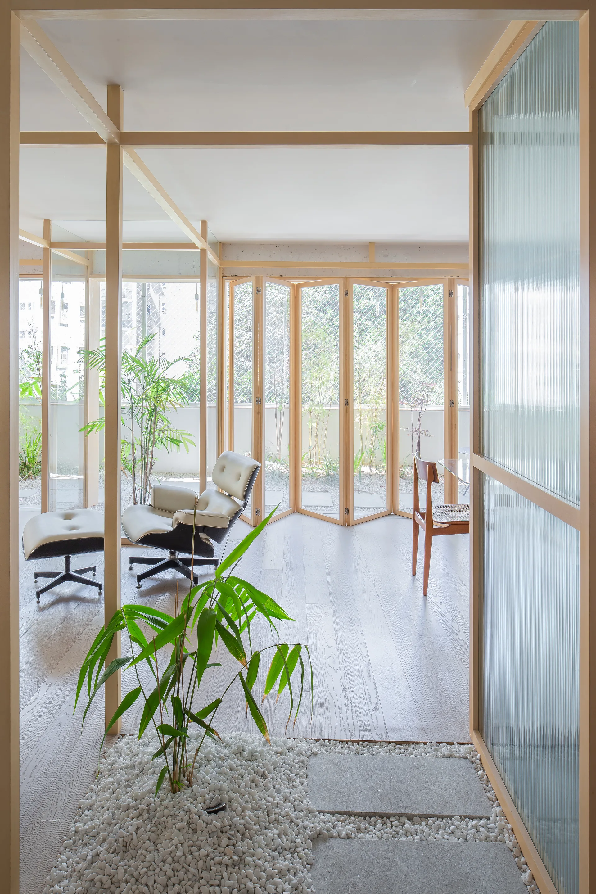 A bright minimalist interior features light wood framing, a ribbed glass partition, a white leather lounge chair, and an indoor pebble garden.