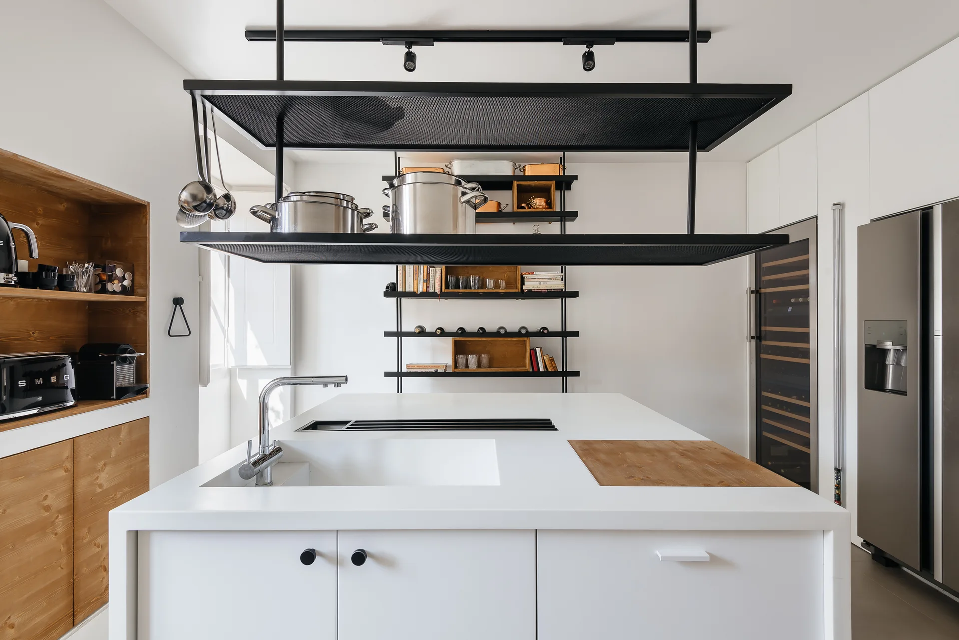 A white minimalist kitchen island with a built-in sink stands beneath a suspended black metal mesh shelving unit.