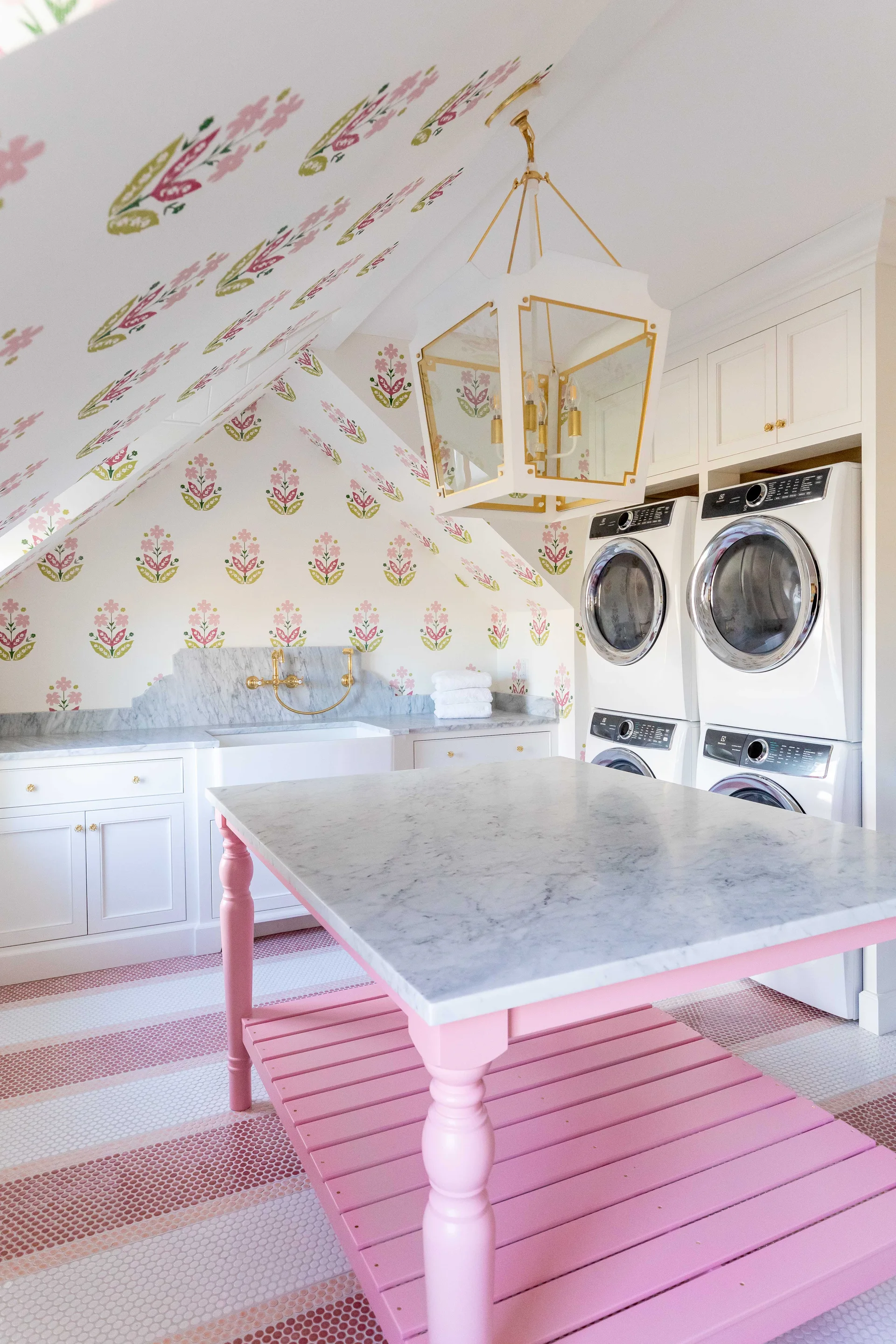 A bright laundry room features floral wallpaper on a sloped ceiling, a pink island with a marble top, and stacked washer-dryer units.