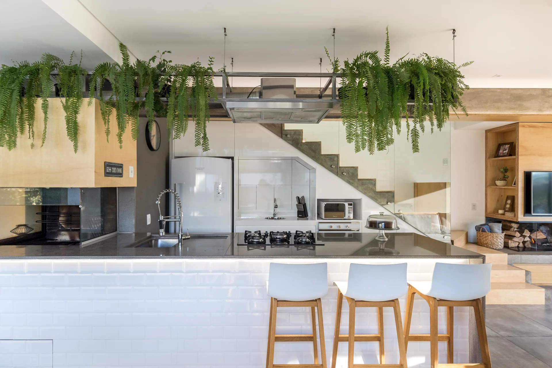A modern kitchen featuring a white subway tile island with bar stools, an integrated cooktop, and ferns hanging from an overhead rack.