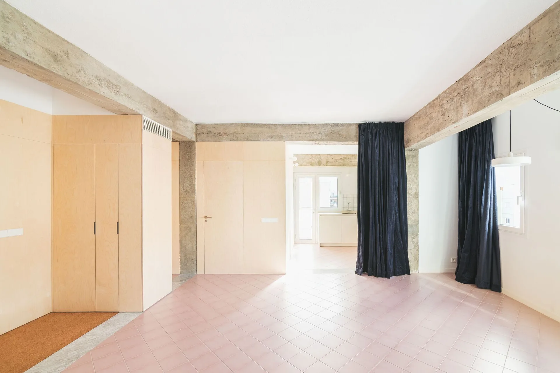 An empty minimalist room features dusty pink floor tiles, light wood wall paneling, exposed concrete beams, and long navy blue curtains.