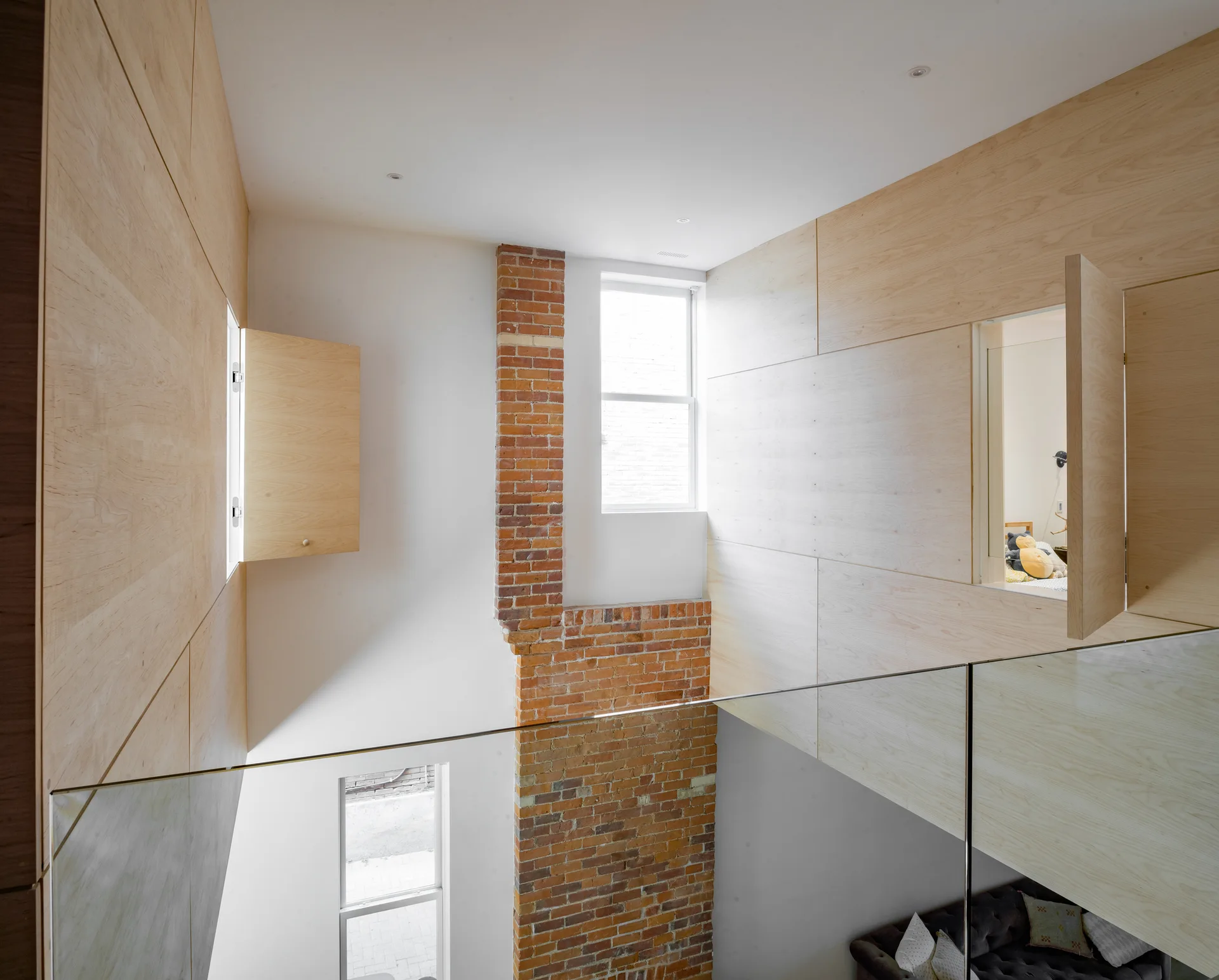 Looking down into a residential stairwell void with light plywood paneled walls, a vertical exposed brick chimney, and a clear glass railing.