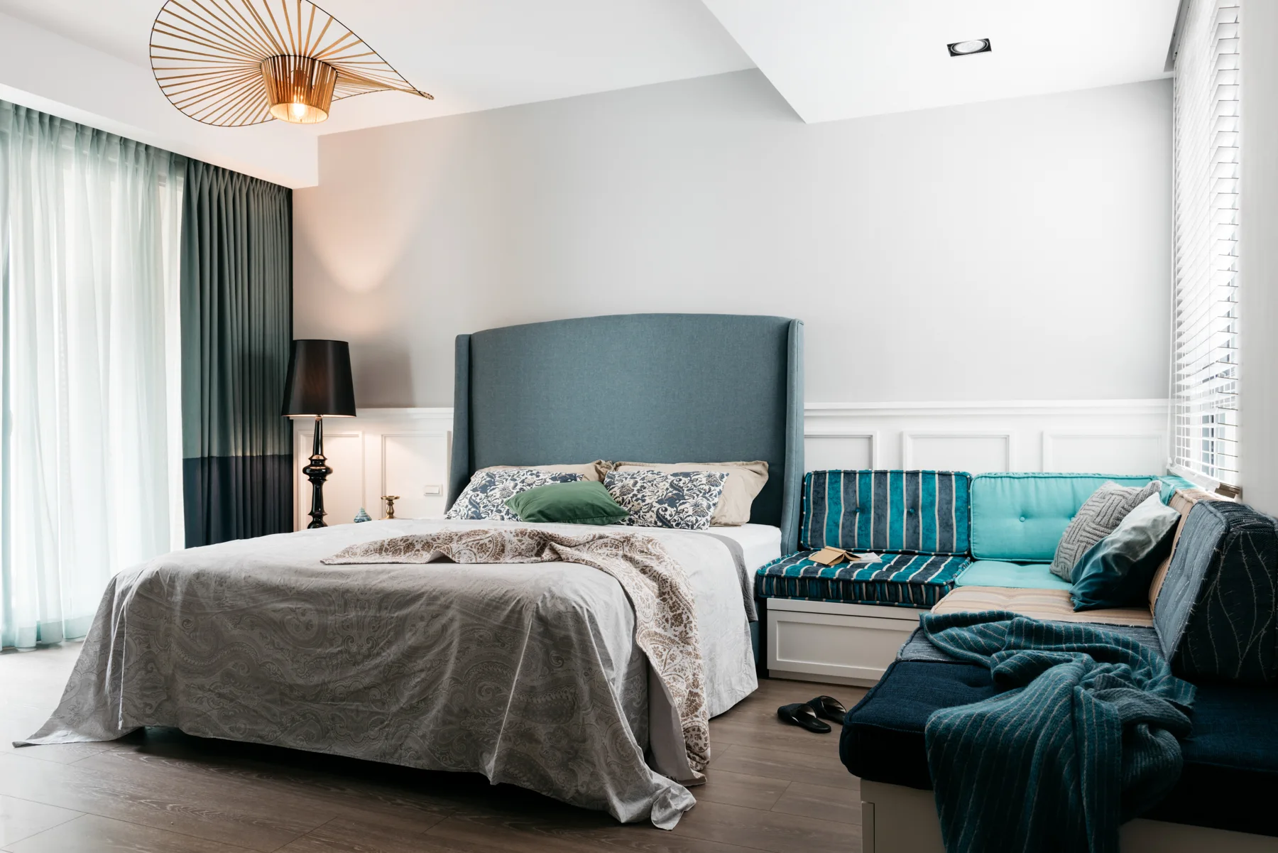 A bedroom featuring a tall blue fabric headboard, a white paneled lower wall, a built-in corner window seat with teal cushions, and a gold wire pendant light.