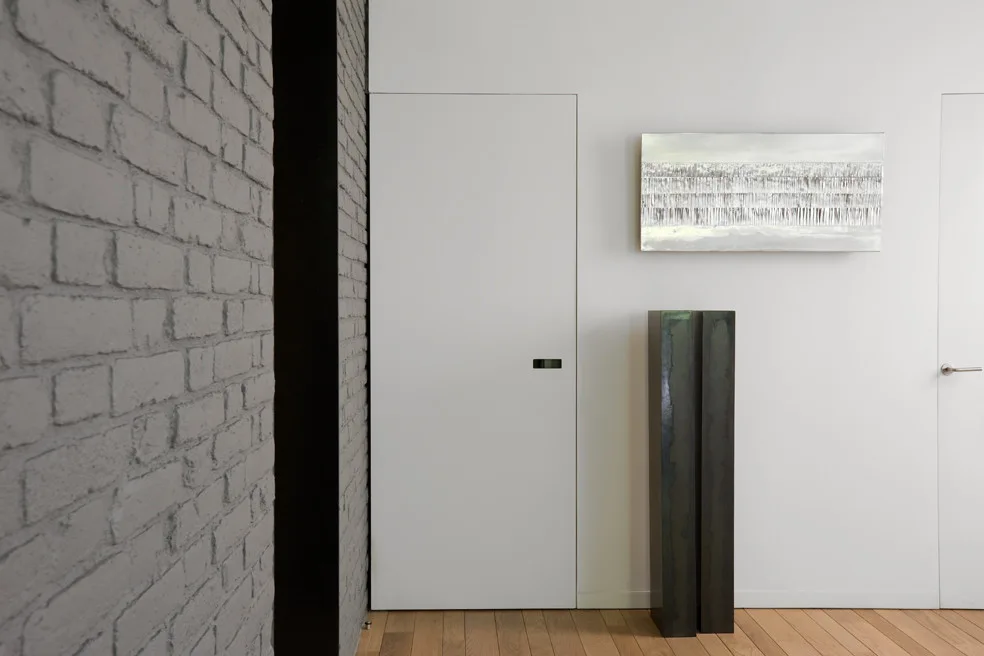 A minimalist hallway features seamless flush white doors, a painted grey brick wall, and a tall dark floor sculpture on light wood floors.