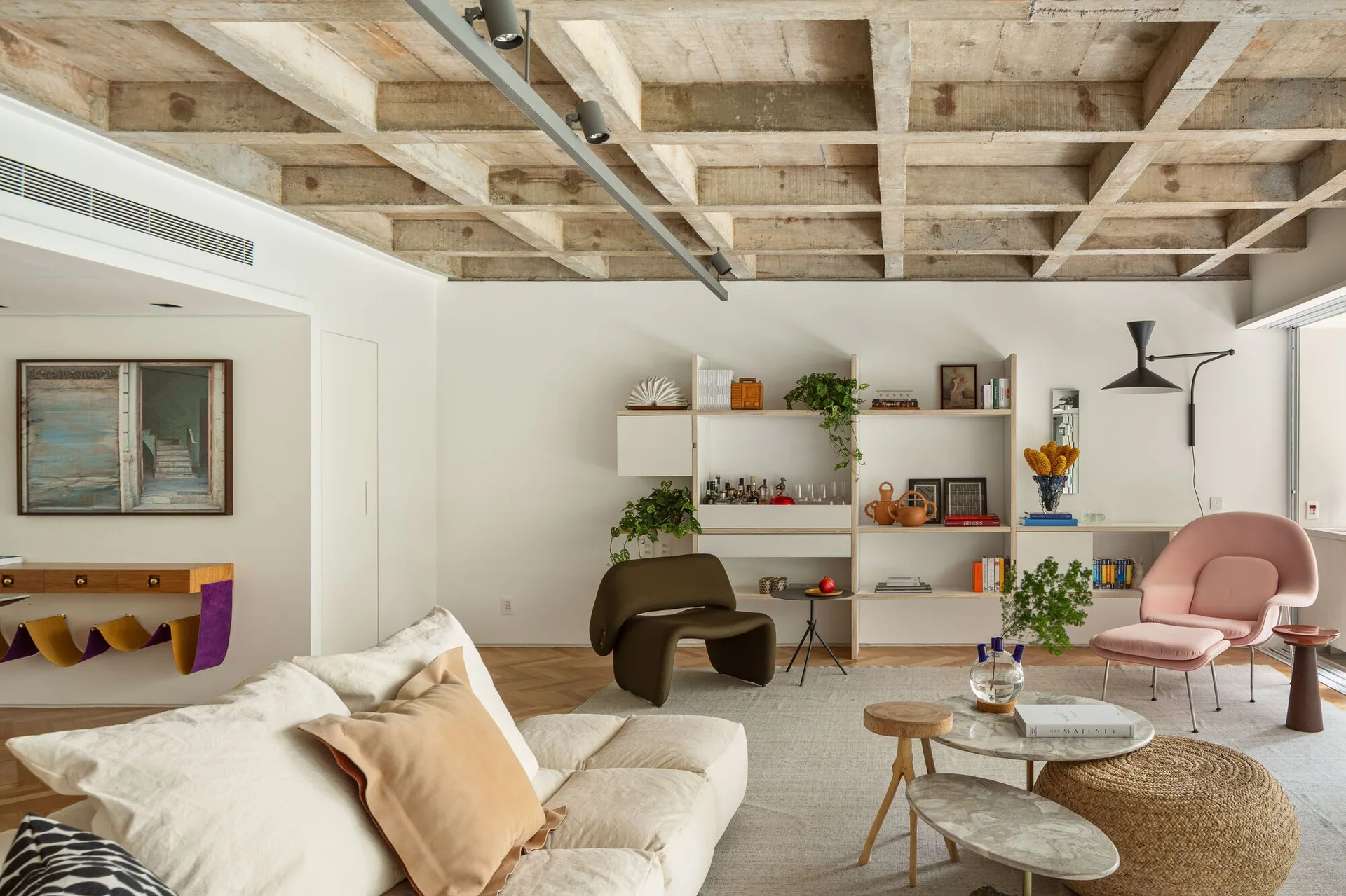 A living room features a cream sofa, tiered marble coffee tables, and a pink lounge chair beneath an exposed concrete waffle ceiling.