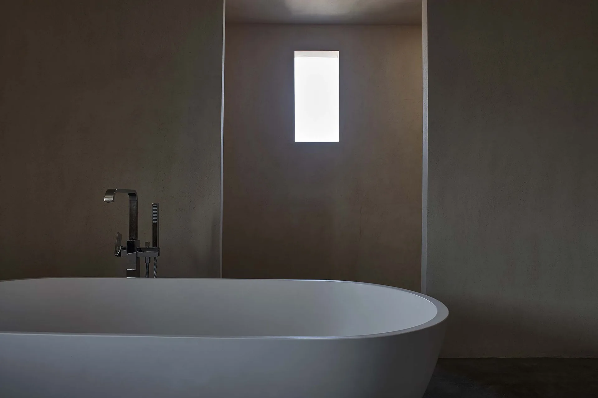 A white freestanding bathtub with a chrome faucet sits in a minimalist bathroom with textured plaster walls and a narrow recessed window.