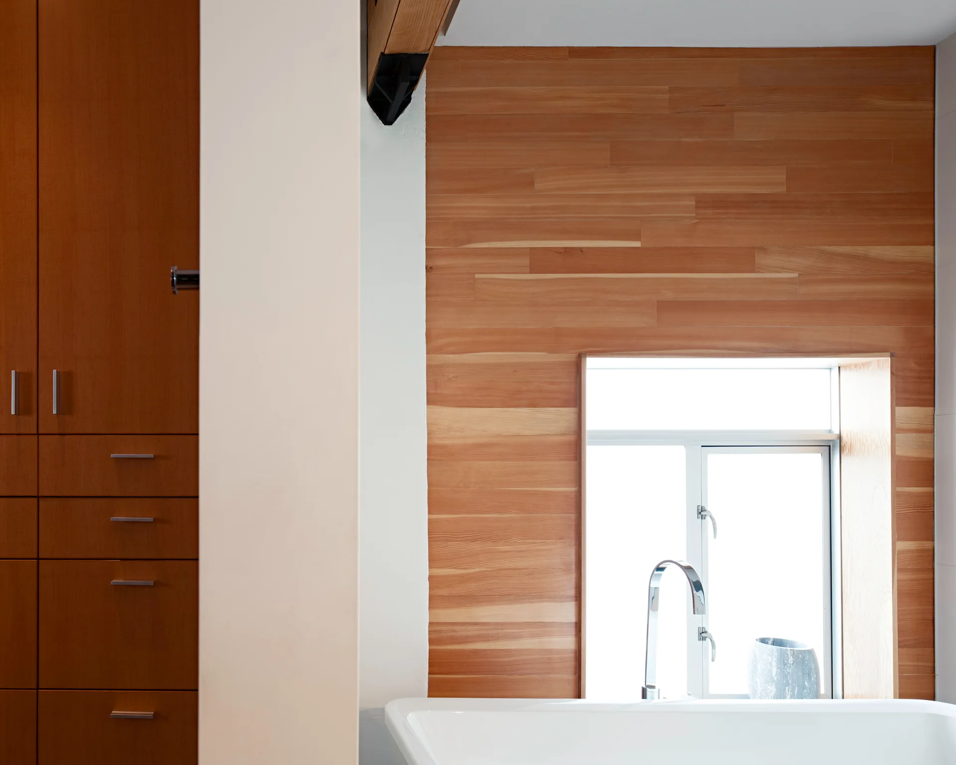 A white bathtub and chrome faucet set against a horizontal wood plank wall with an inset window and adjacent brown cabinetry.