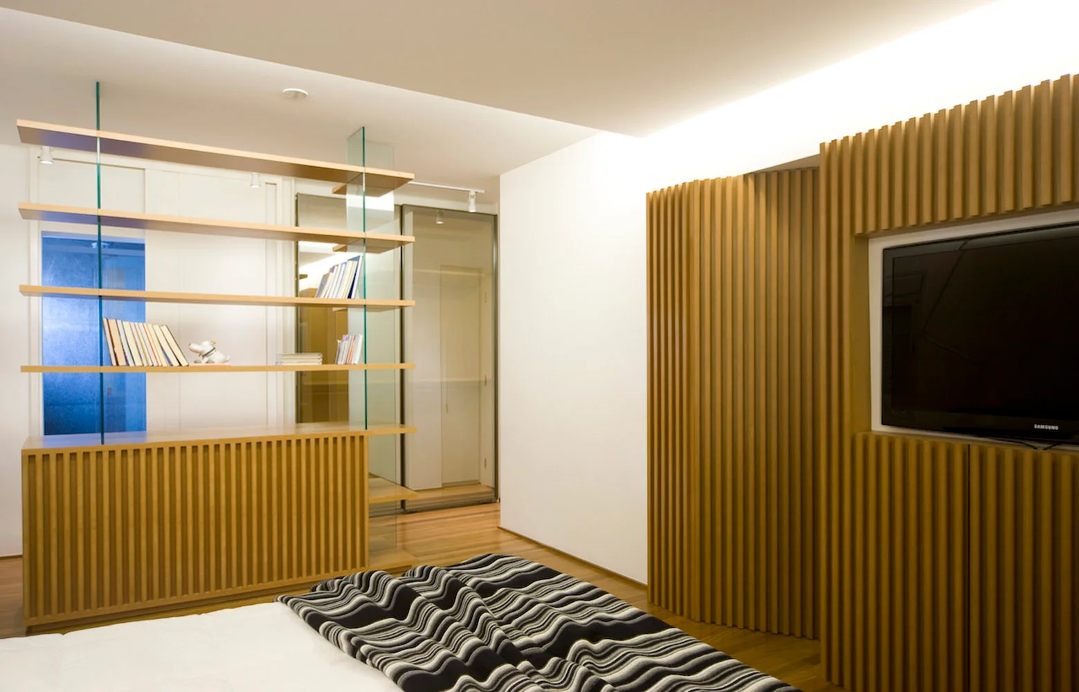 A modern bedroom featuring a bed with a striped throw, a custom wooden slatted wall panel with a TV, and open shelves with glass supports.