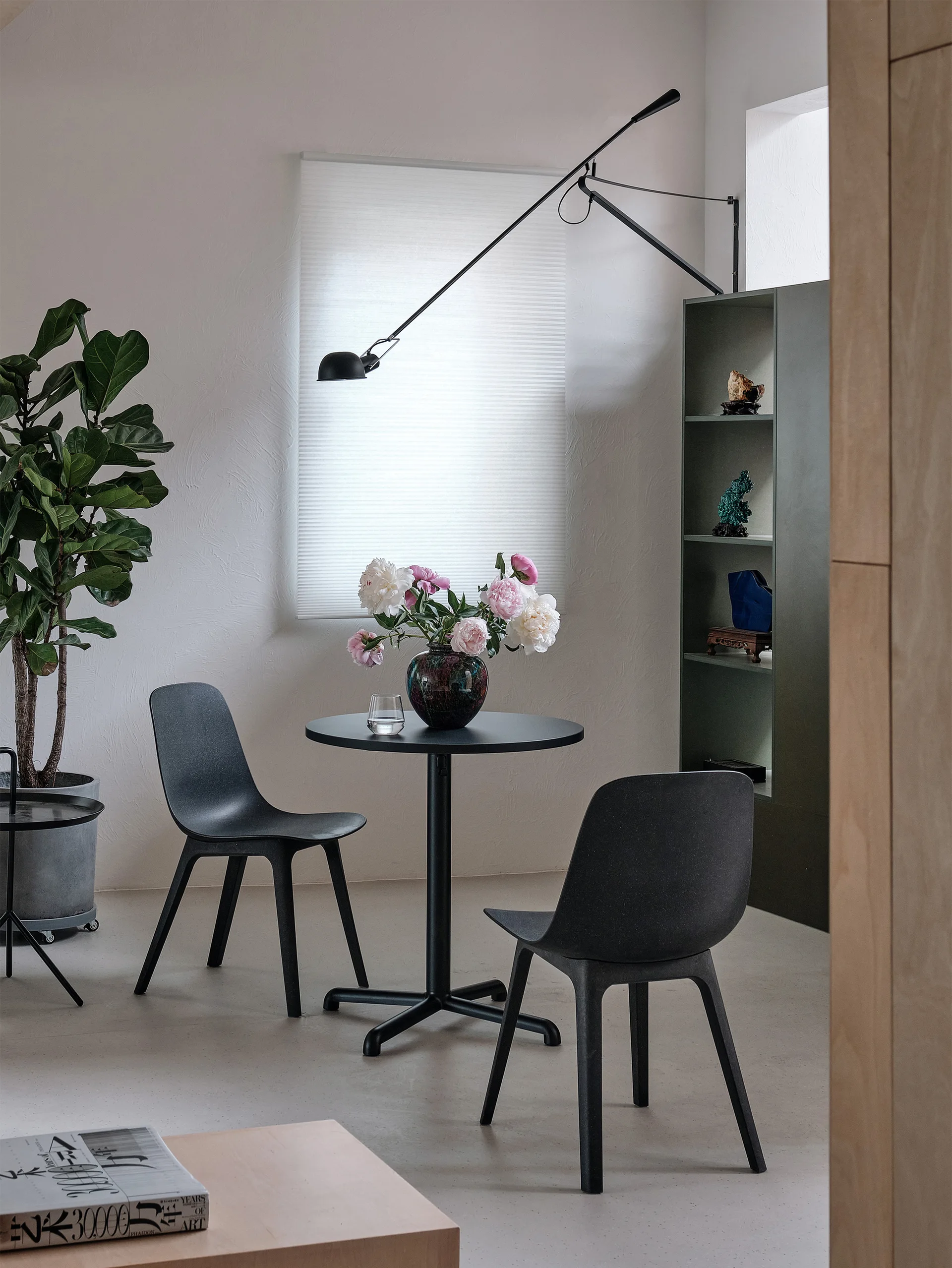 A minimalist dining nook features a black bistro table with two chairs, an articulated wall lamp, and a green shelving unit.