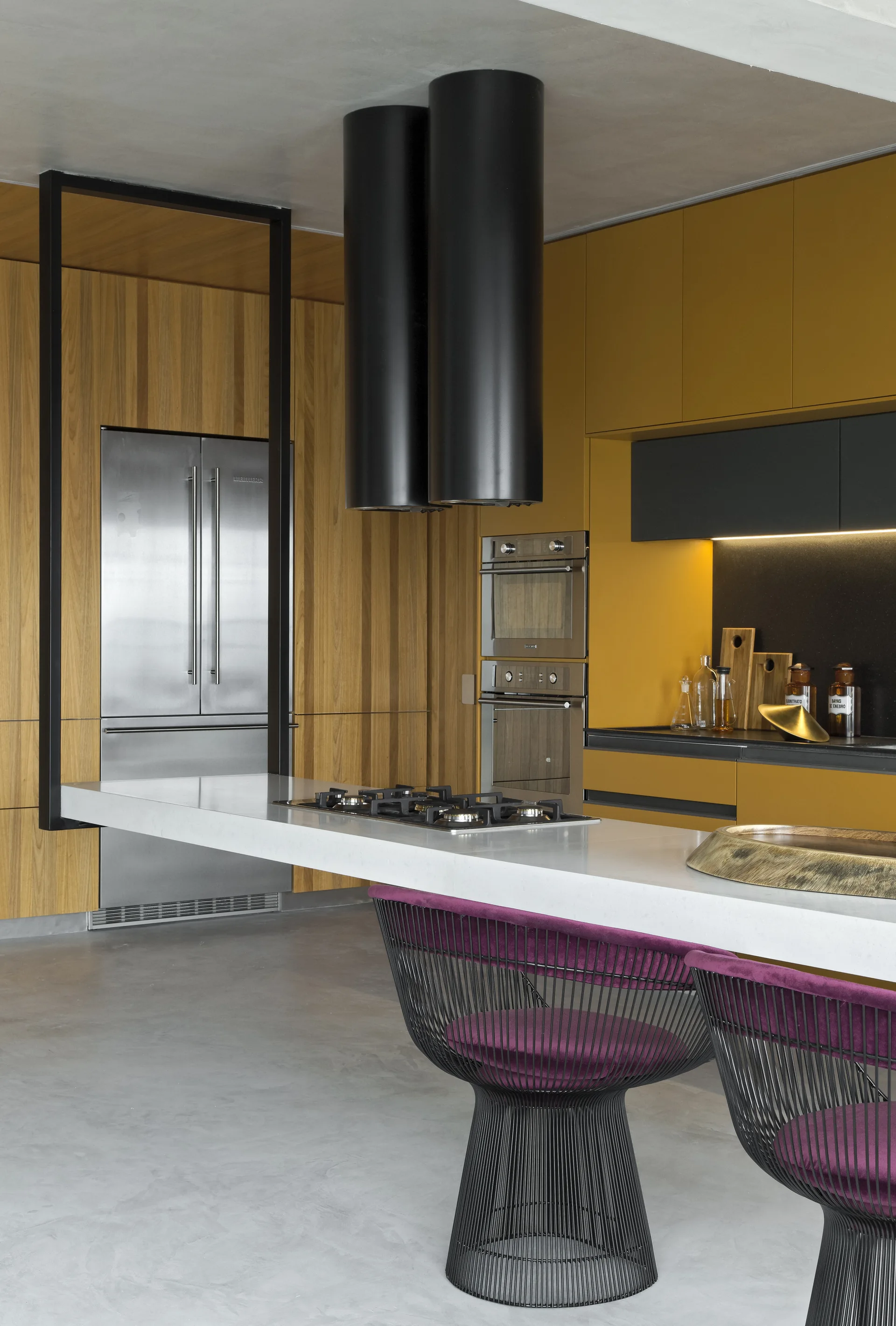 A modern kitchen features mustard yellow cabinetry, a white island with black cylindrical range hoods, and purple wire stools on a concrete floor.