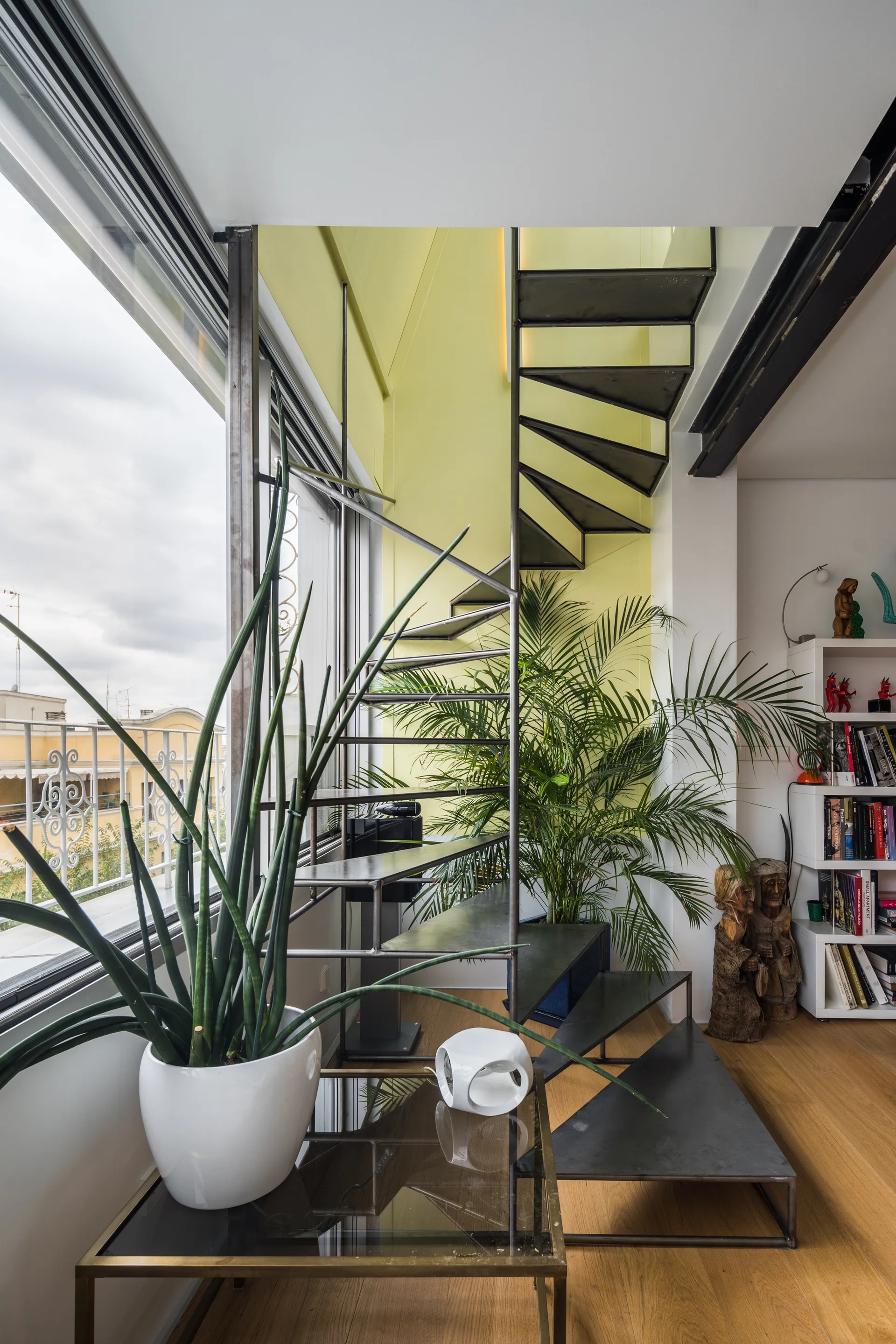 A black metal spiral staircase ascends beside a large window and a pale yellow wall, with potted plants on light wood floors.