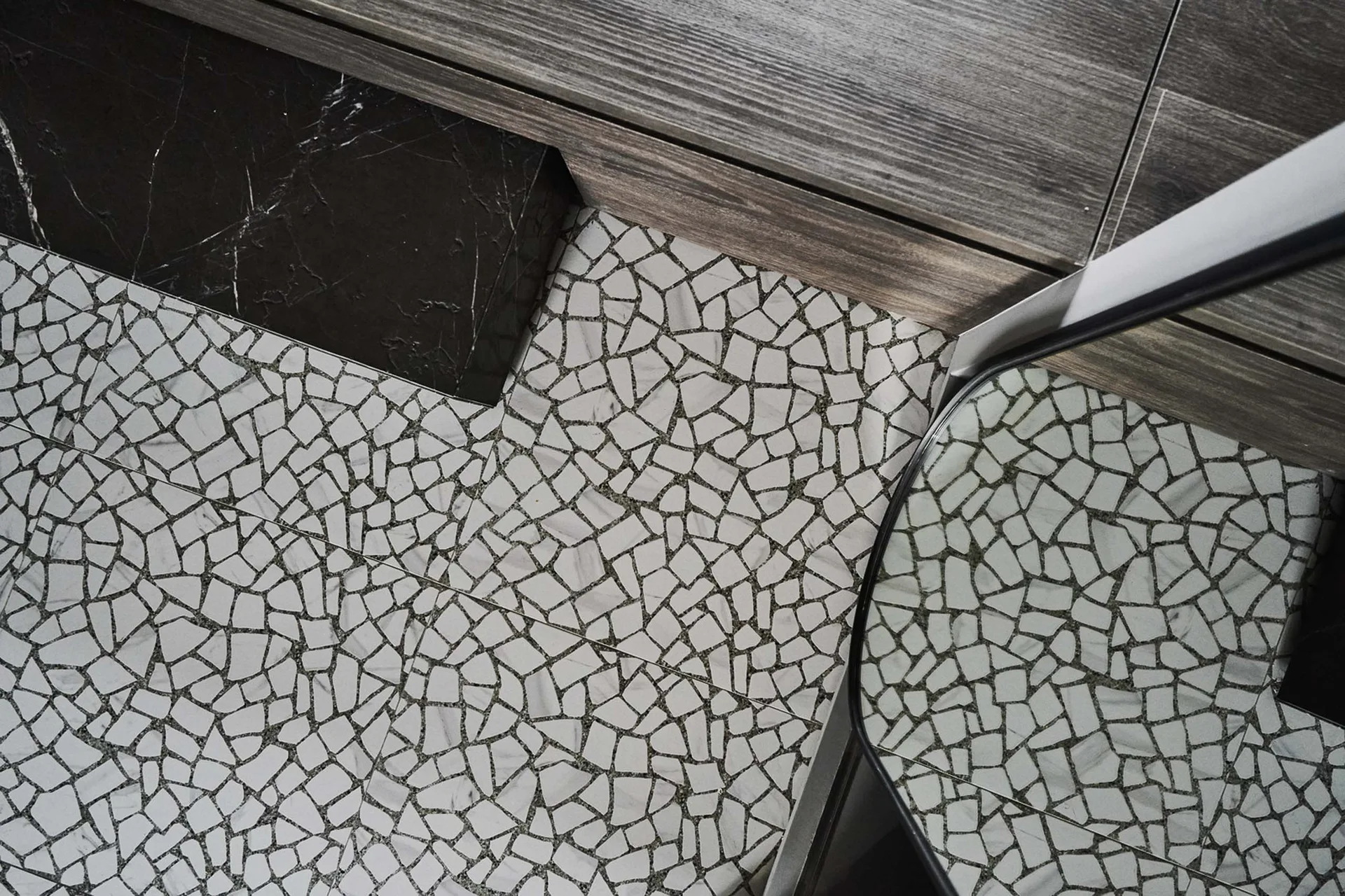 An overhead view of an irregular geometric stone mosaic tile floor meeting a grey wood-plank surface and a polished black marble block.