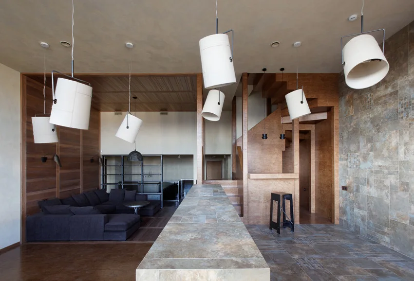 A split-level room features a stone island in the foreground, a charcoal sectional sofa in a wood-paneled area, and angled white cylinder pendant lights.