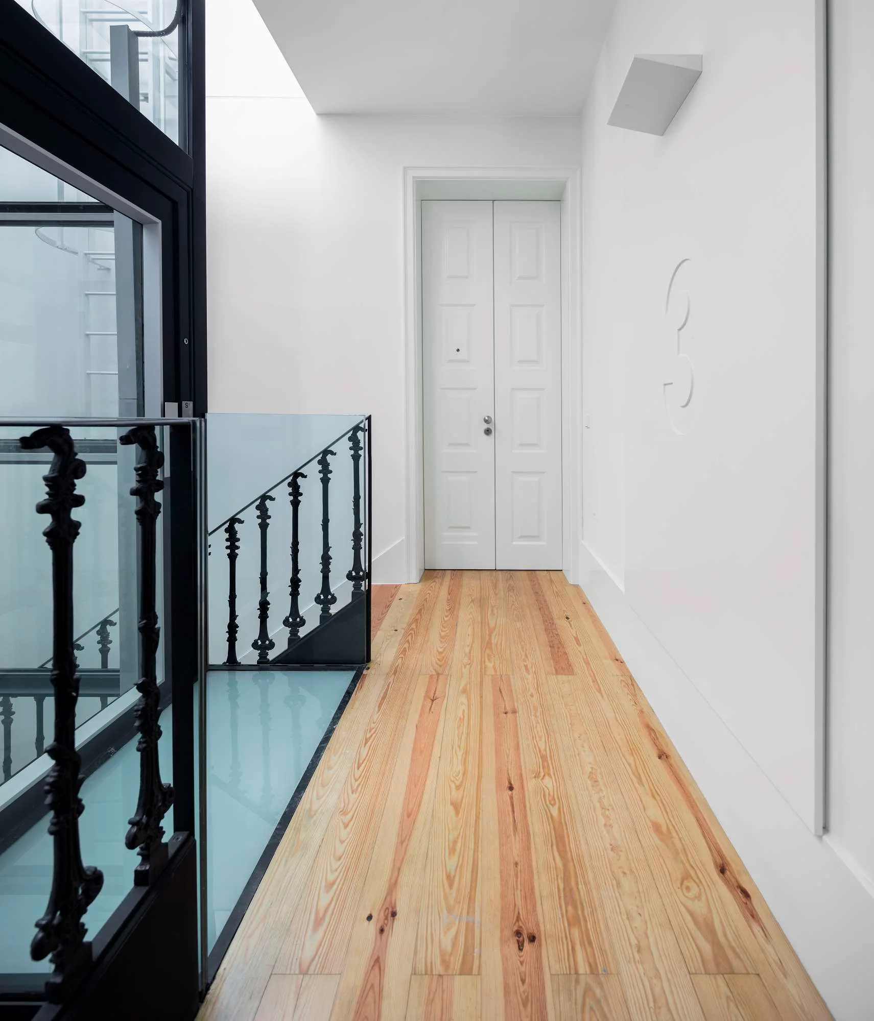 A bright white hallway with natural wood plank flooring, a white paneled door, and an ornate black metal staircase railing.