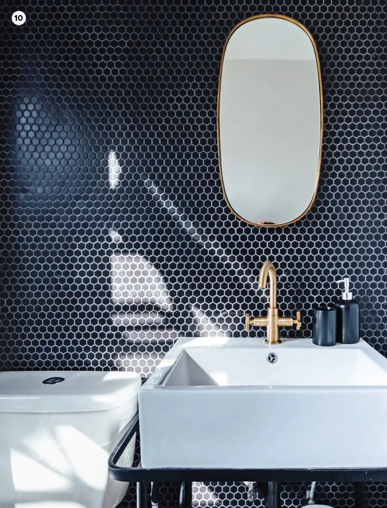 A bathroom featuring a white ceramic sink with a brass faucet, an oval gold-framed mirror, and a wall covered in black hexagonal tiles.