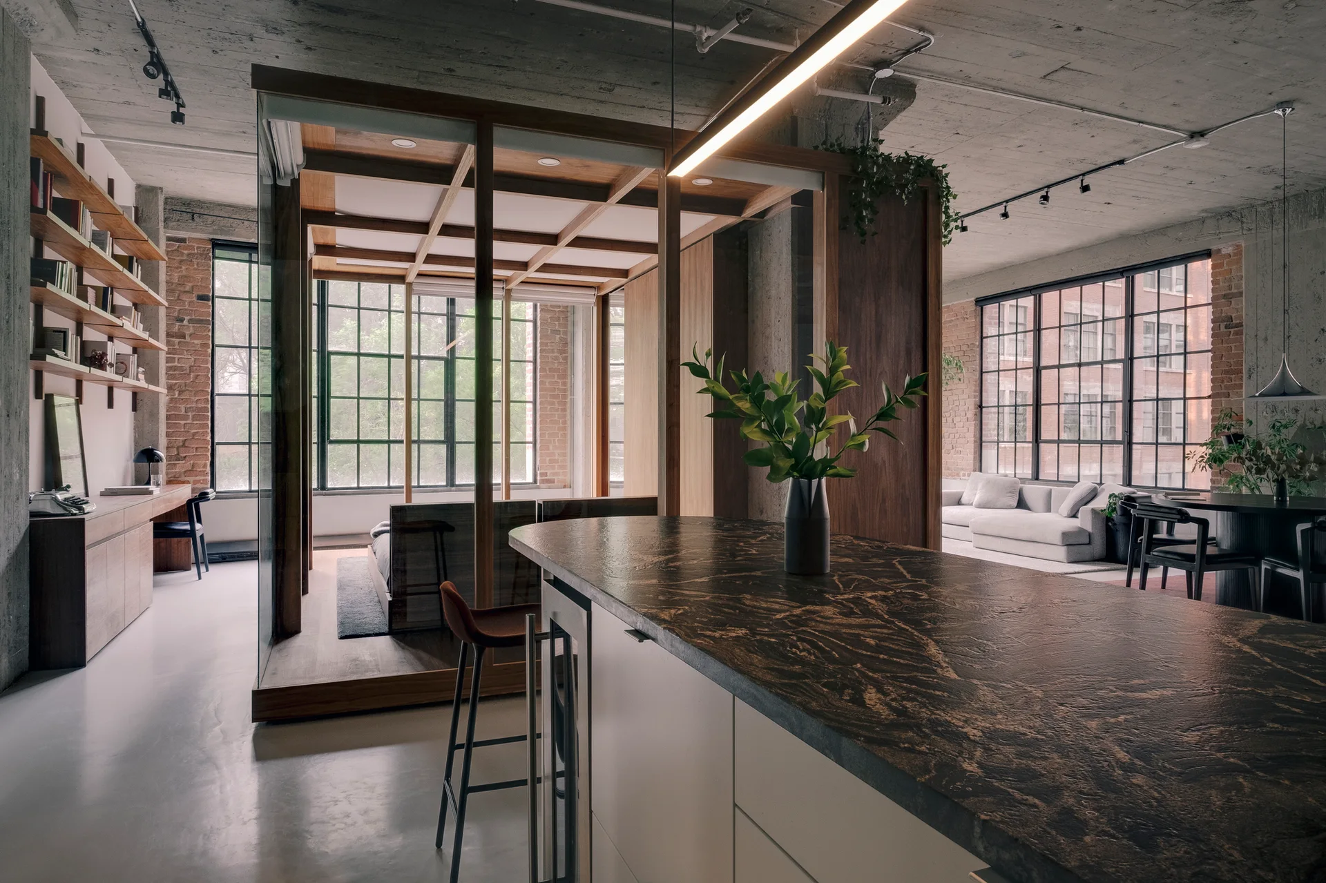 An industrial loft featuring a dark stone kitchen island in the foreground and a glass-enclosed bedroom structure framed in wood in the background.