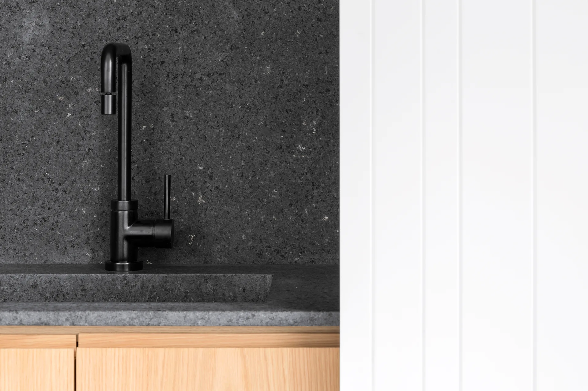 A matte black faucet stands against a dark stone backsplash and integrated sink, paired with light wood cabinets and a white slatted wall.