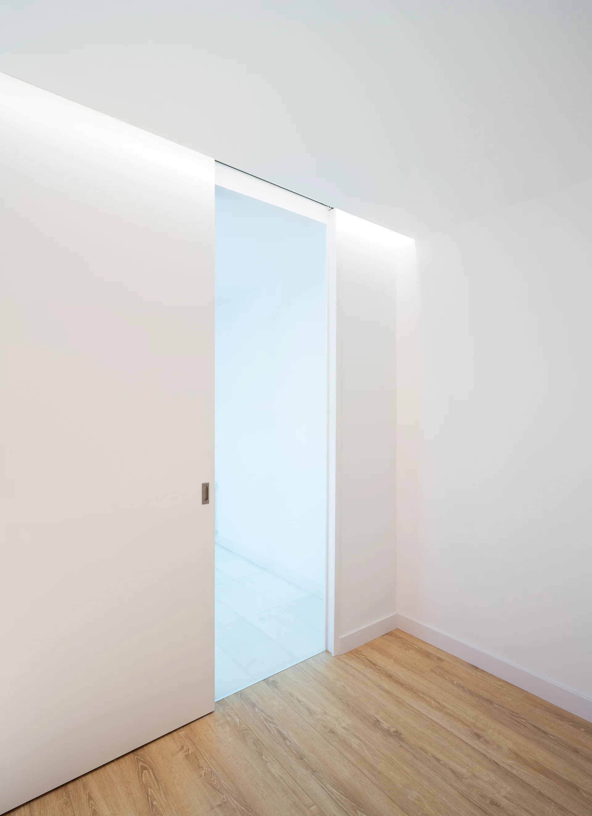 A minimalist white sliding pocket door stands partially open on light wood floors beneath a recessed cove light.