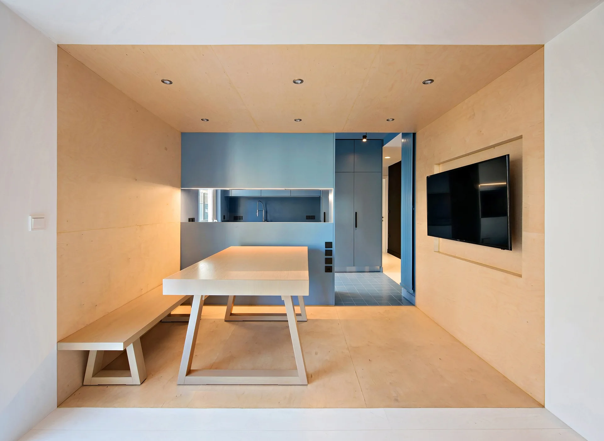 A modern dining area features a light wood table and bench enclosed in plywood paneling, with a blue kitchen alcove in the background.