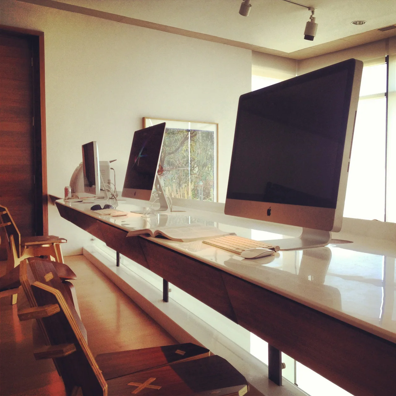 A long minimalist wooden desk with a white glossy top holds multiple desktop computers accompanied by modern wooden chairs in a bright workspace.