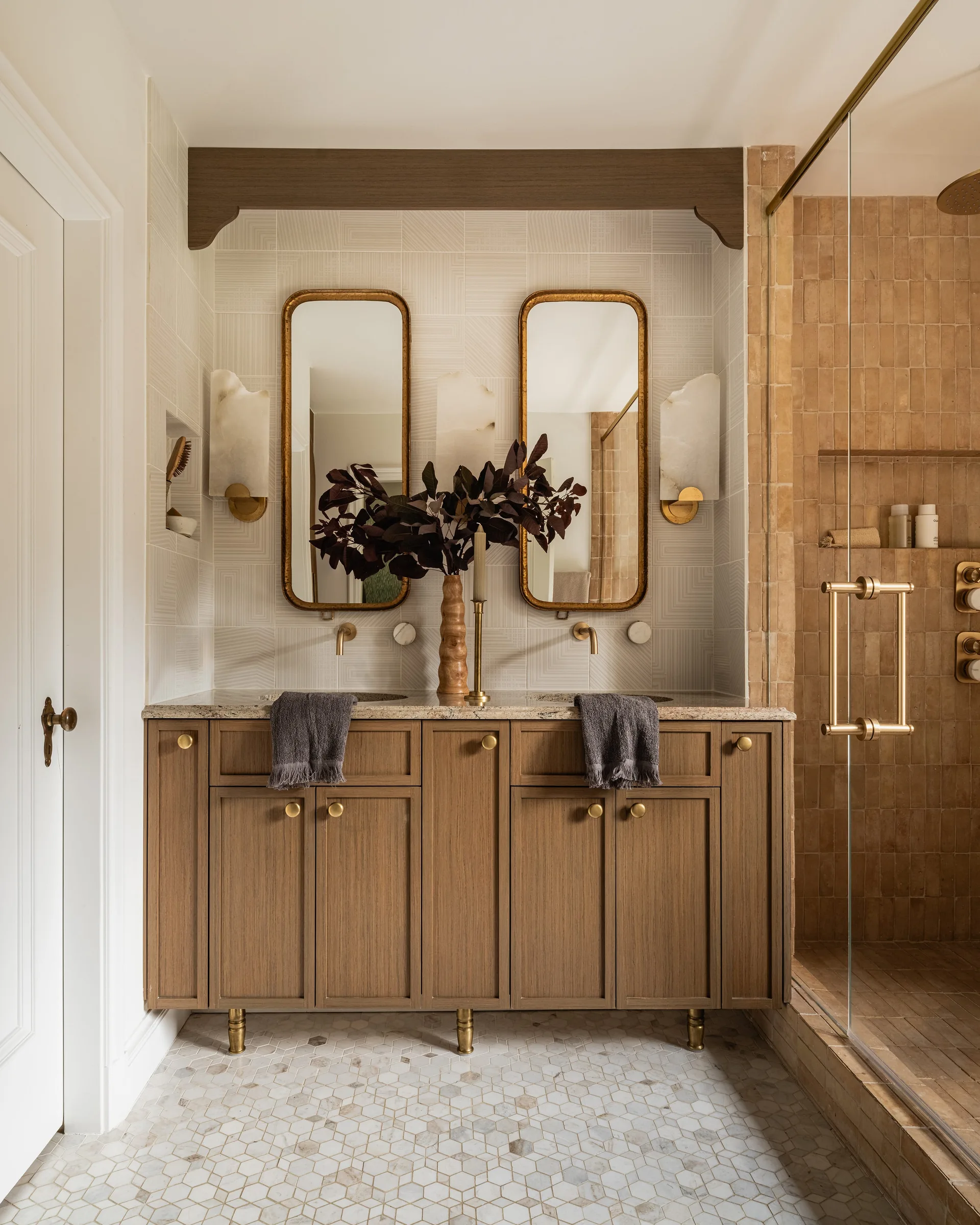 A double bathroom vanity features a natural wood cabinet with gold hardware, twin rounded brass mirrors, stone wall sconces, and hexagon floor tiles.