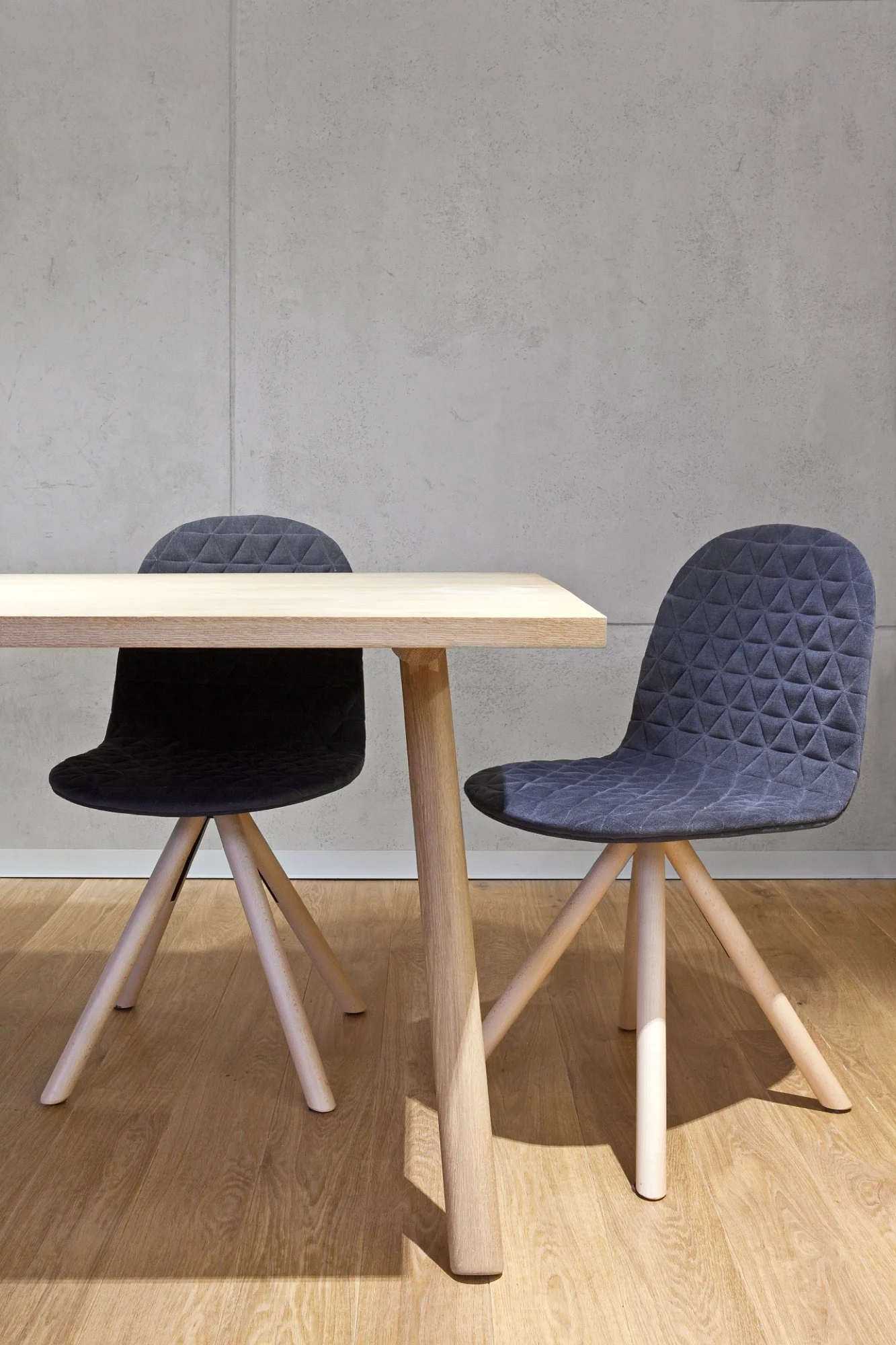 A light wood dining table and two charcoal gray upholstered chairs with quilted backs stand on a wood floor against a concrete wall.
